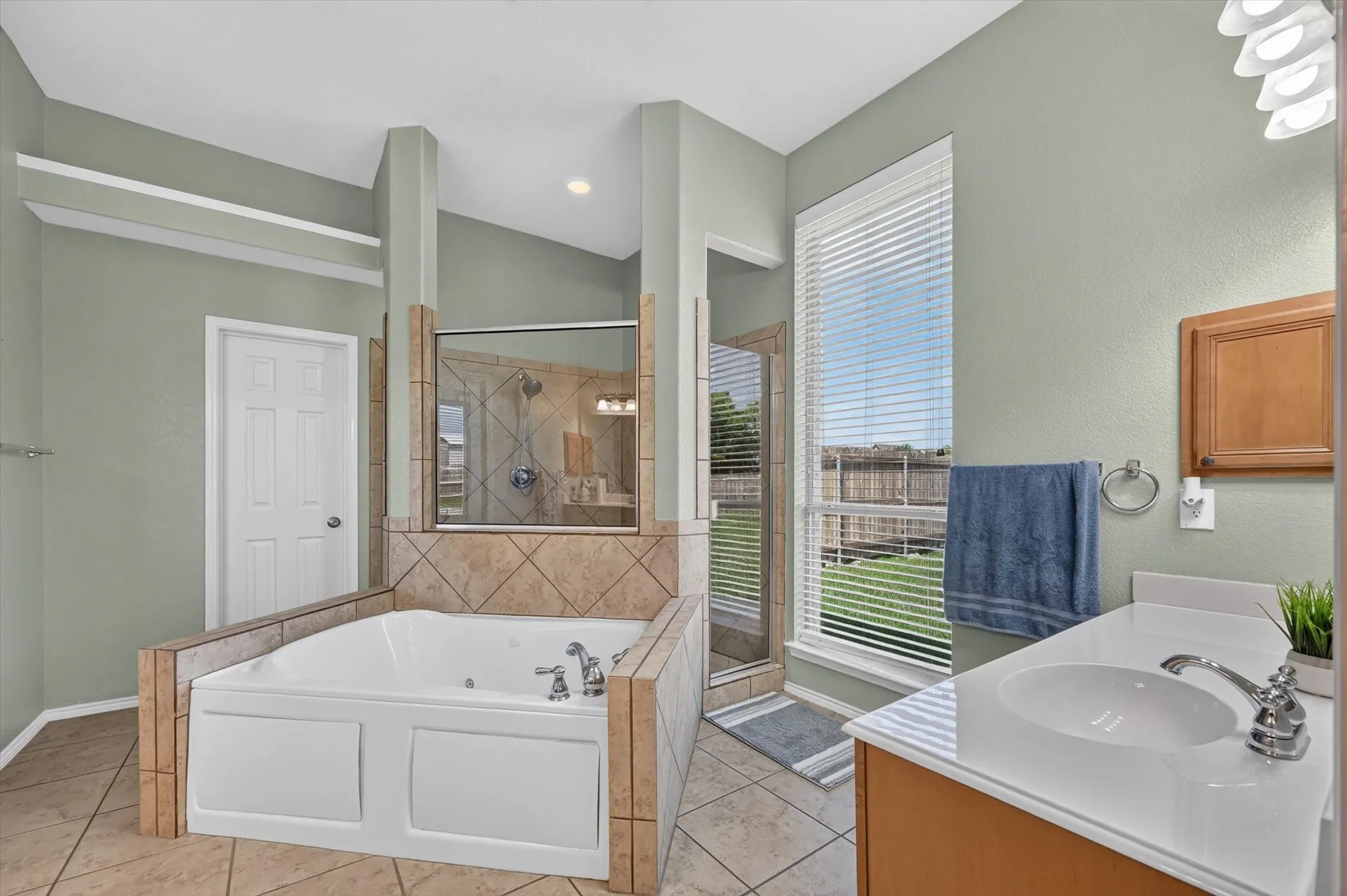 Bathroom featuring a built-in jetted tub with tile surround, a separate glass-enclosed shower, a single vanity with an integrated sink and laminate countertop, a wood-finish wall cabinet, and a large window with blinds