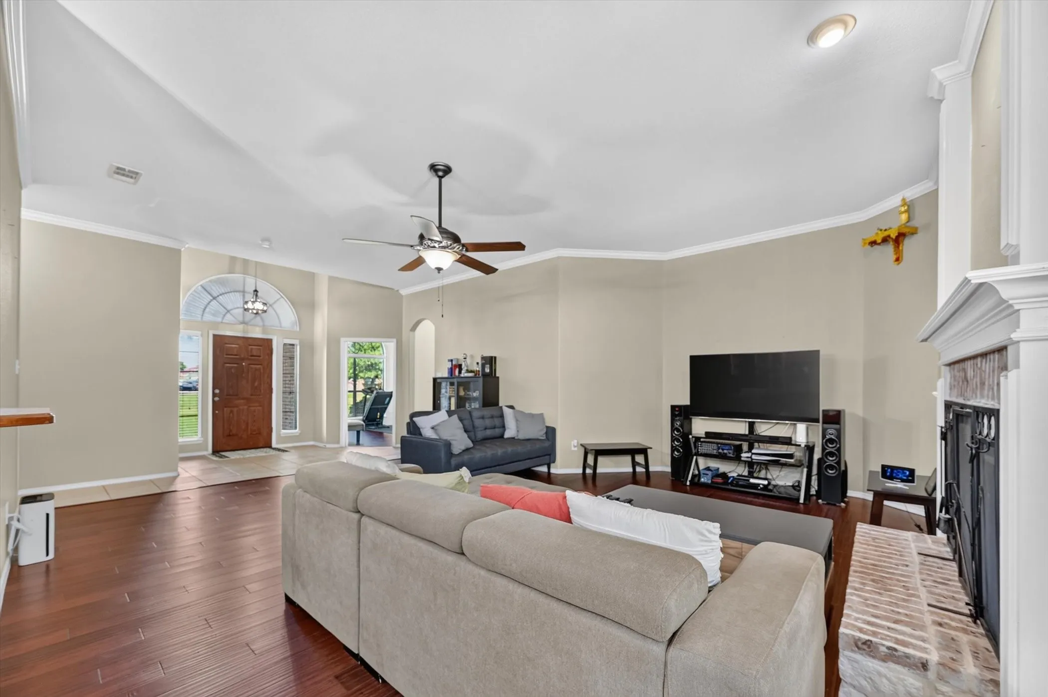 Spacious living area featuring wood-finish flooring, a ceiling fan, crown molding, and a prominent fireplace with a brick surround and white mantel