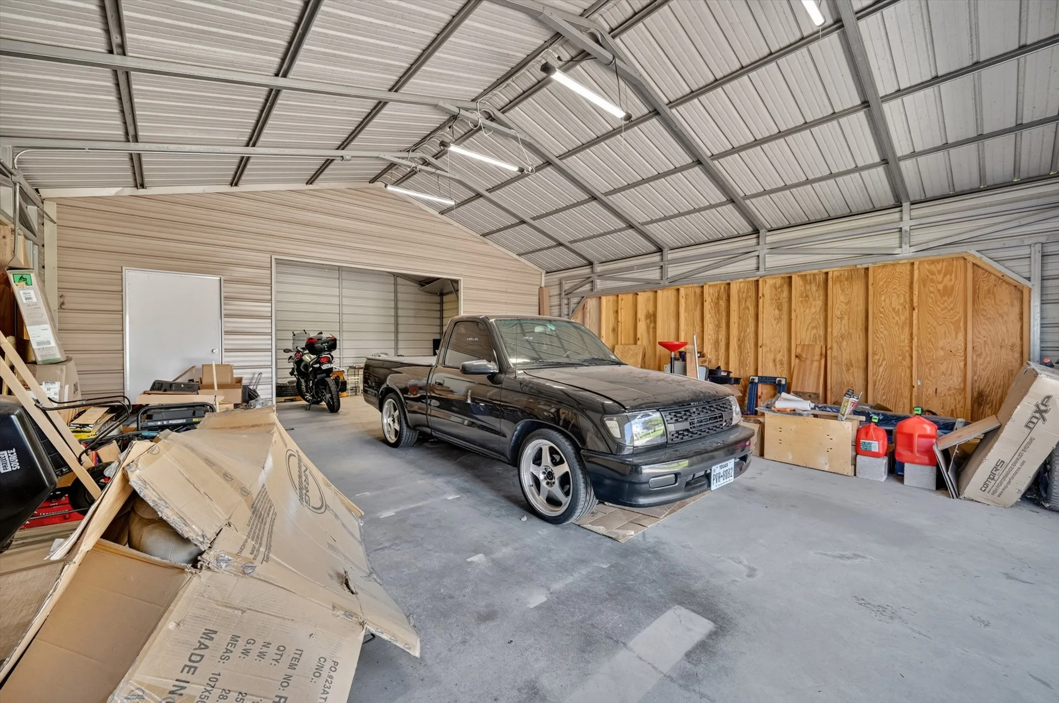 Spacious metal garage structure featuring a concrete floor, a corrugated metal roof with visible trusses, and integrated fluorescent lighting