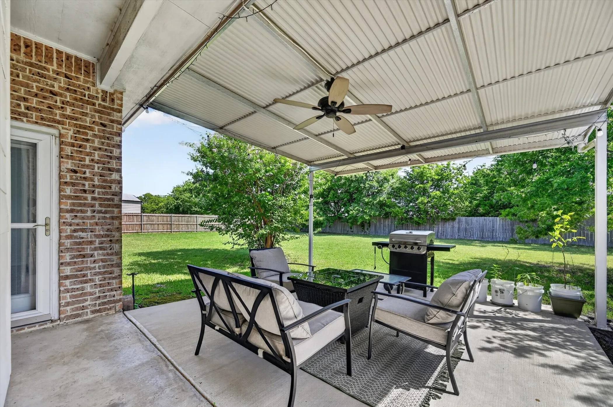Covered patio featuring a ceiling fan, brick exterior, concrete slab, and expansive grassy yard with mature trees