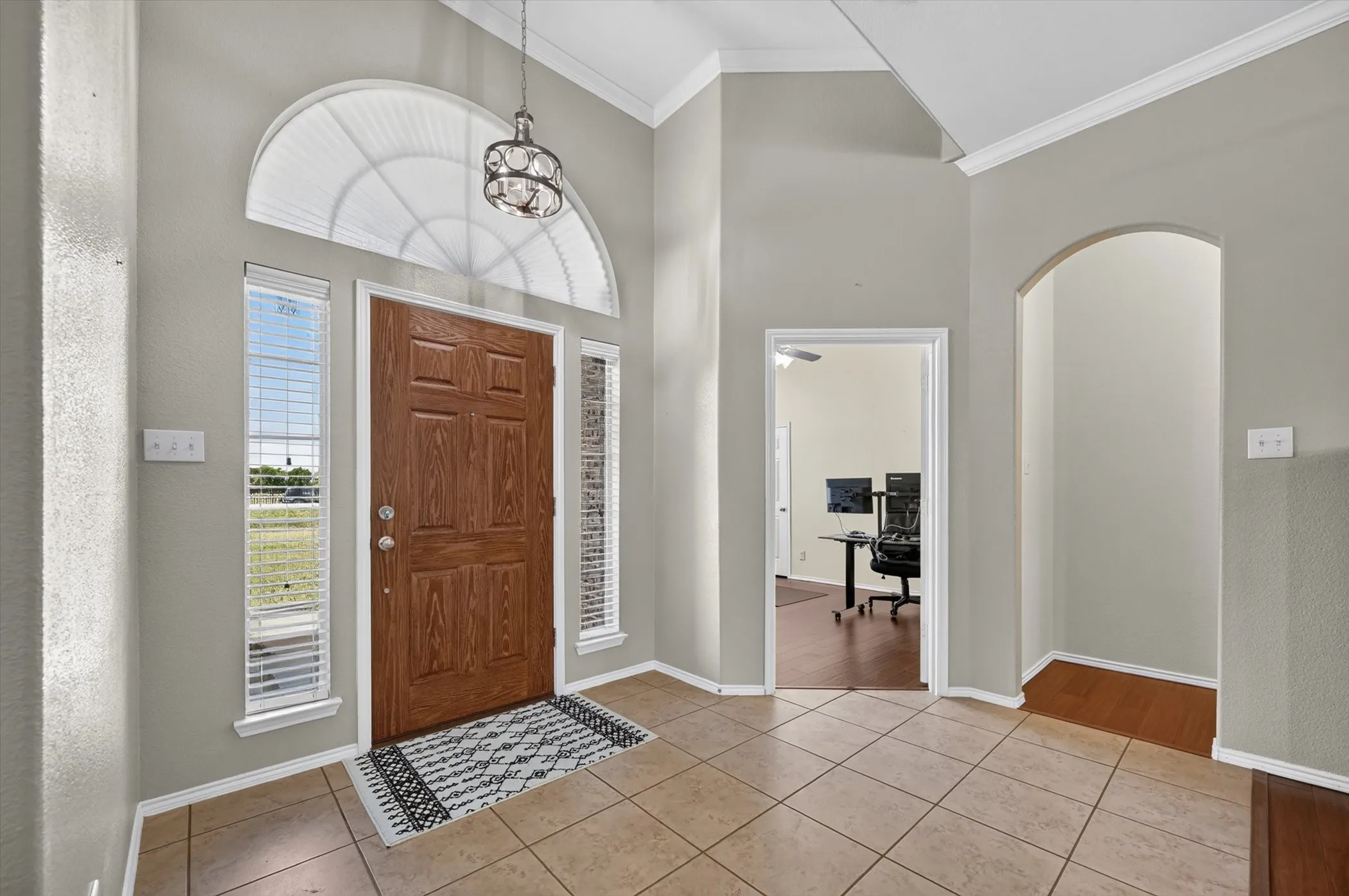 Entryway featuring a paneled wood door with sidelights and an arched transom window, a decorative hanging light fixture, vaulted ceilings with crown molding, and tile flooring