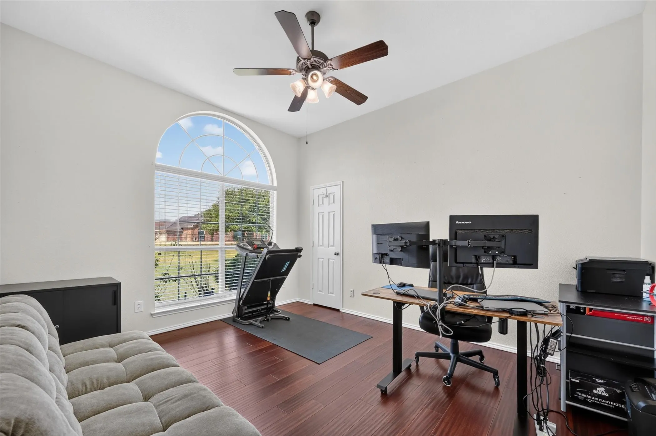 Room featuring a large arched window with blinds, dark wood-finish flooring, a ceiling fan with integrated lighting, a white painted door, and light-colored walls