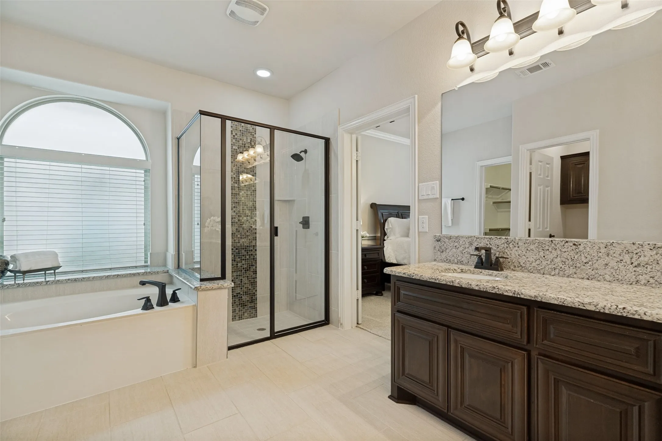 Bright bathroom featuring a large soaking tub with a black faucet, a framed glass shower with mosaic tile, a dual vanity with a light stone countertop, and light tile flooring