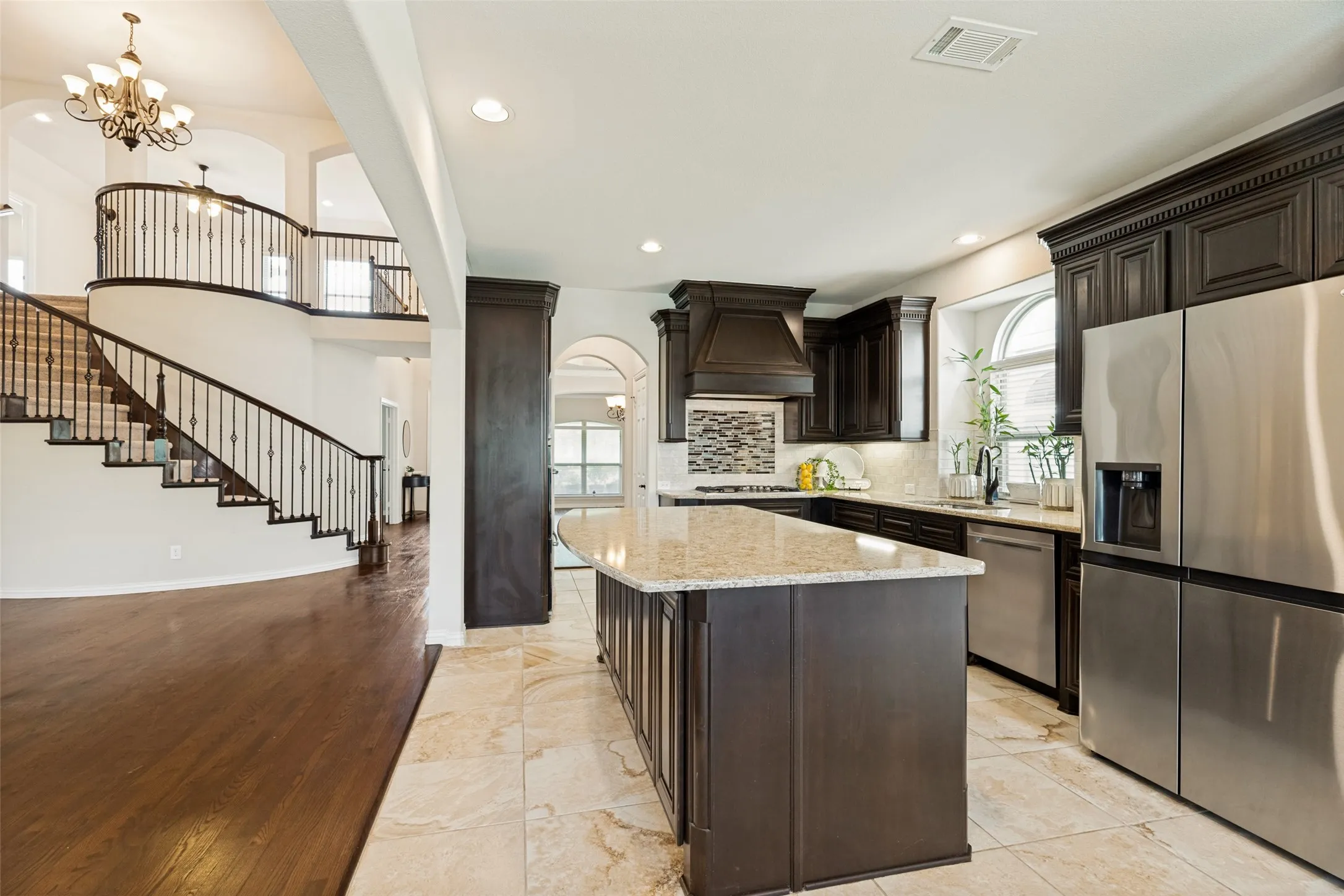 Gourmet kitchen featuring dark wood cabinetry, stainless steel appliances, and an island with a light granite-style countertop, opening to a grand staircase with wrought iron balusters and wood-finish flooring