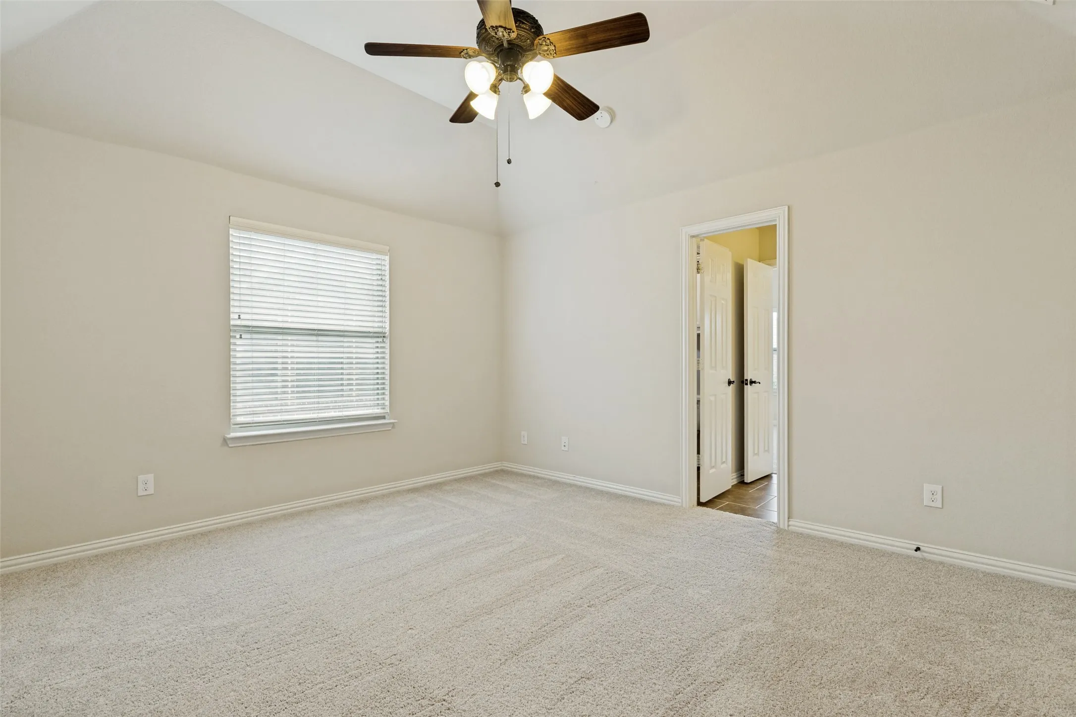 Spacious room featuring vaulted ceilings, neutral-toned carpet flooring, a window with blinds, and a ceiling fan with integrated lighting