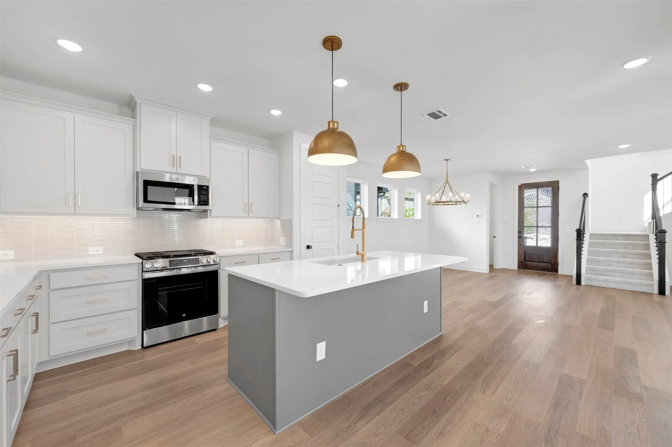 Bright kitchen with white cabinetry, stainless steel appliances, a center island with sink, wood-finish flooring, and a dark wood entry door