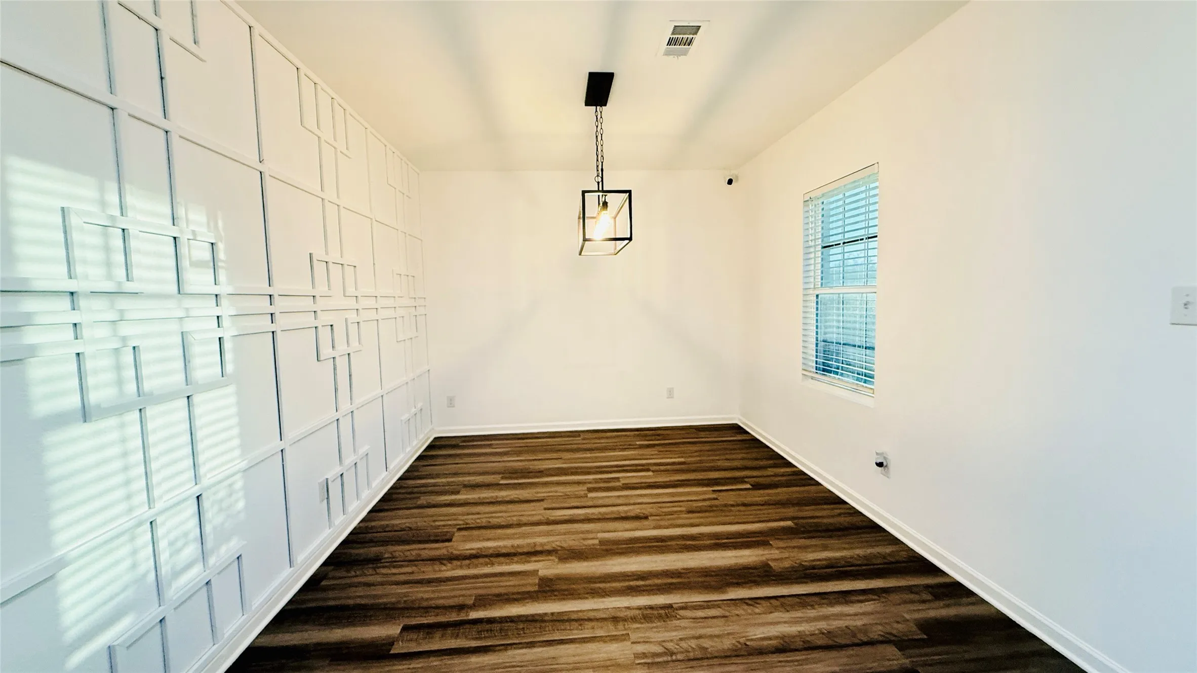 Room featuring an accent wall with geometric molding, laminate-finish flooring, a contemporary black metal pendant light fixture, and a window with blinds