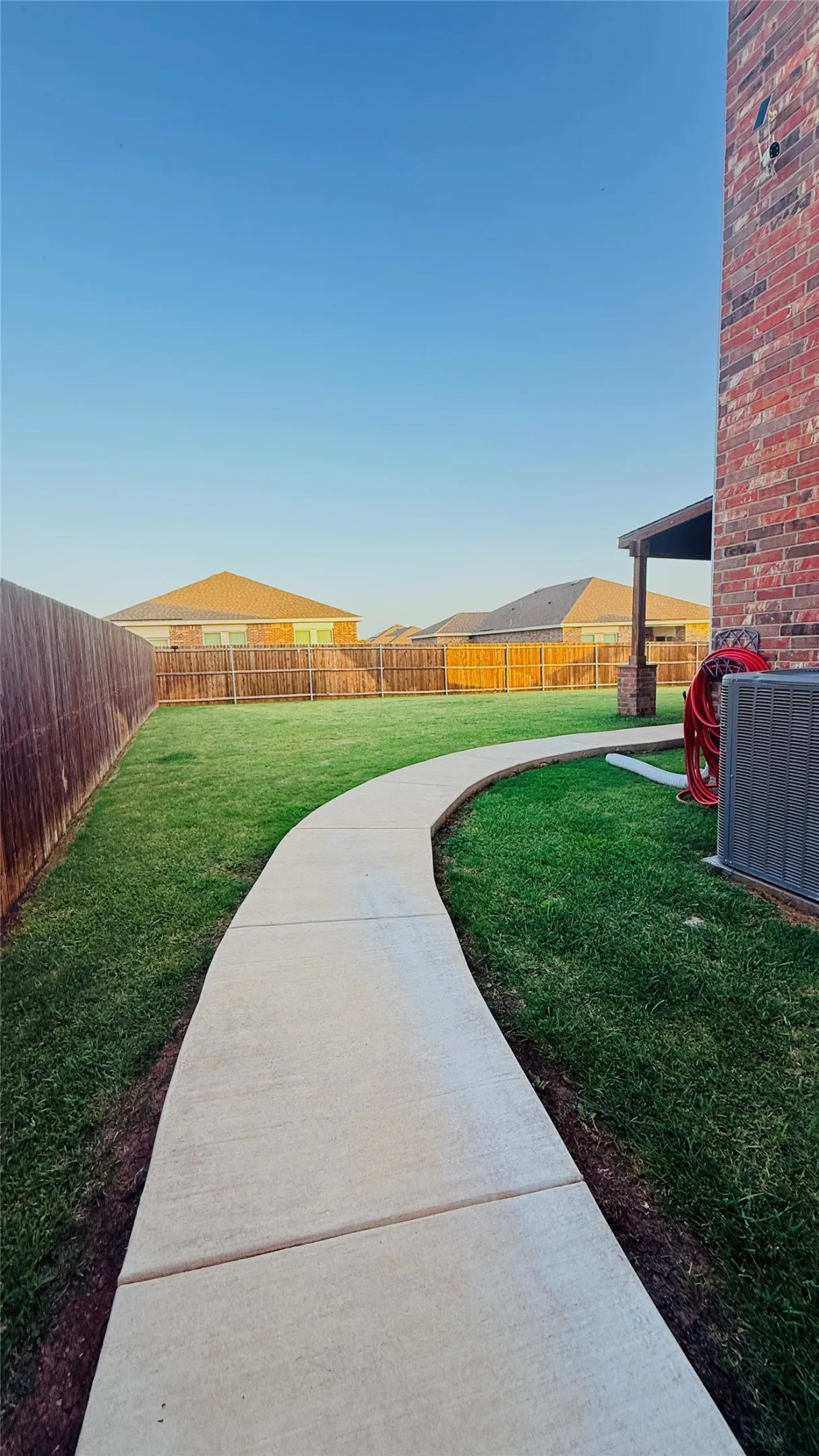 Curved concrete pathway bordering a lush lawn, featuring a privacy fence, brick exterior, and covered patio