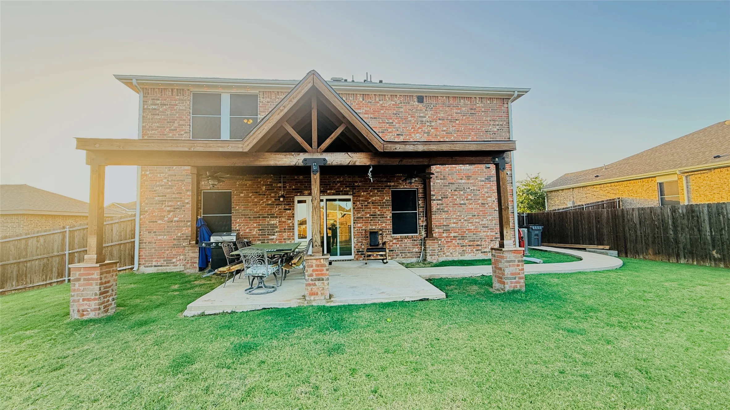 Expansive backyard featuring a covered patio with exposed wood beams, brick columns, and a concrete slab