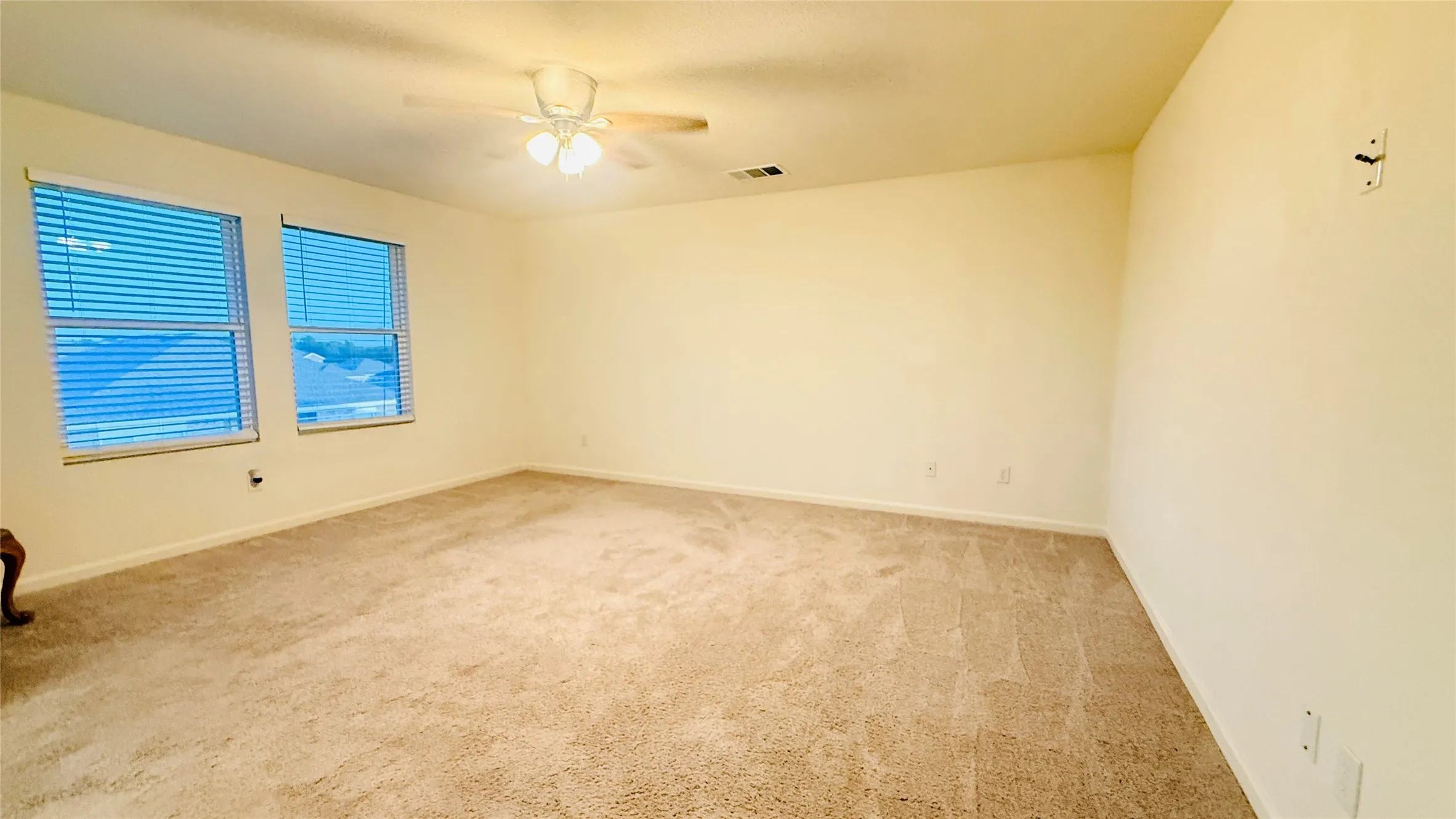 Spacious room featuring neutral carpeting, two large windows with blinds, a ceiling fan with integrated lighting, and light-toned wall finishes