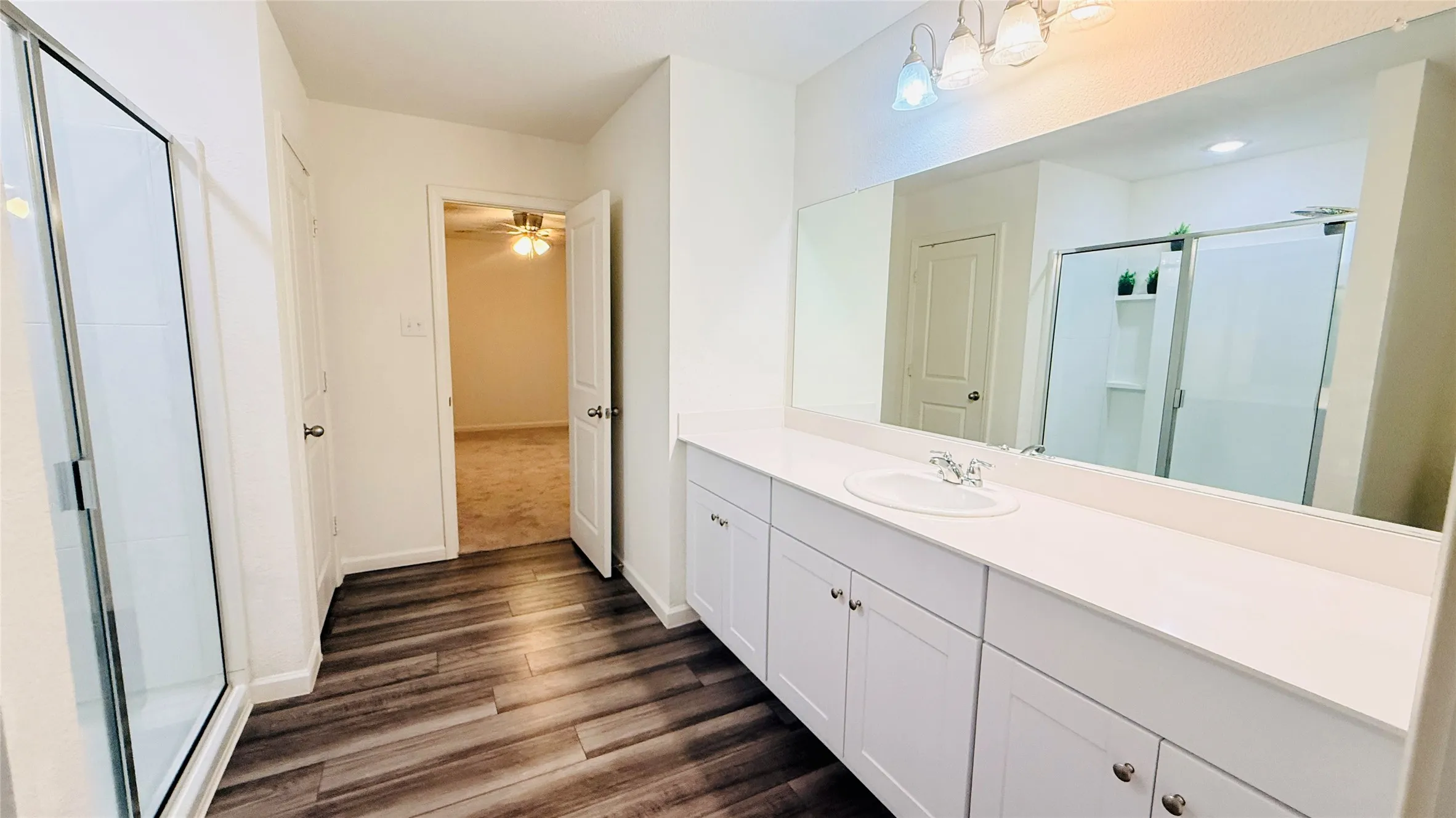 Full bathroom featuring Laminate-finish flooring, a glass-enclosed shower, a long vanity with dual under-mount sinks, white cabinetry, and a large wall-mounted mirror
