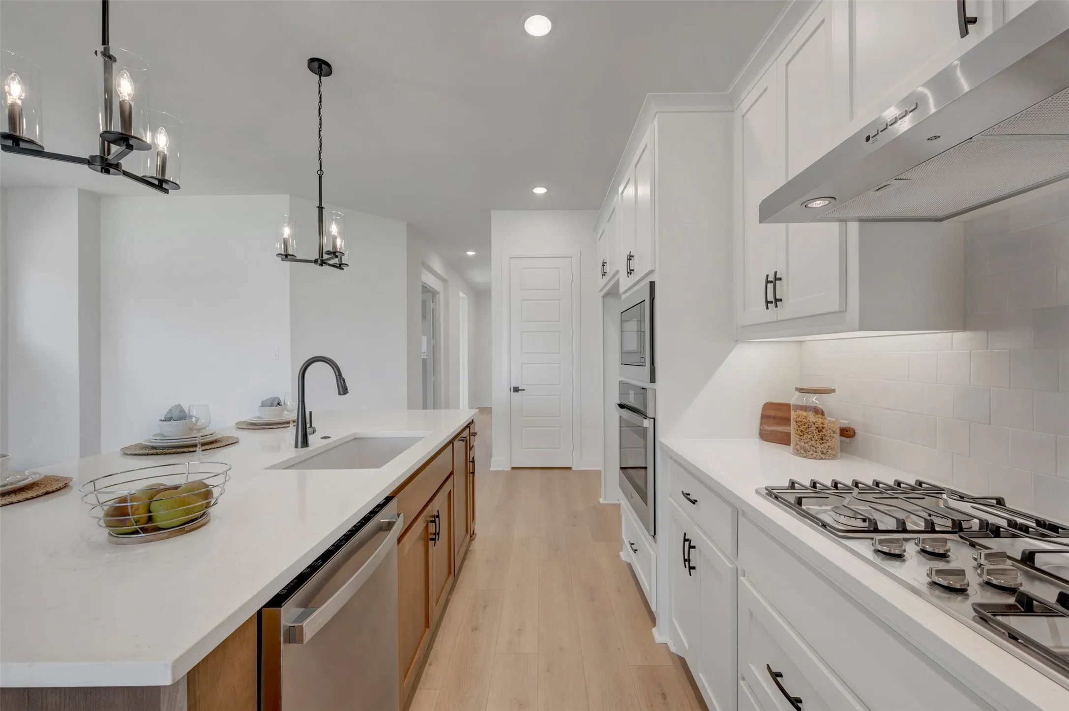 Spacious kitchen featuring an expansive island with an undermount sink, stainless steel appliances, white subway tile backsplash, white shaker cabinetry, and wood-finish flooring