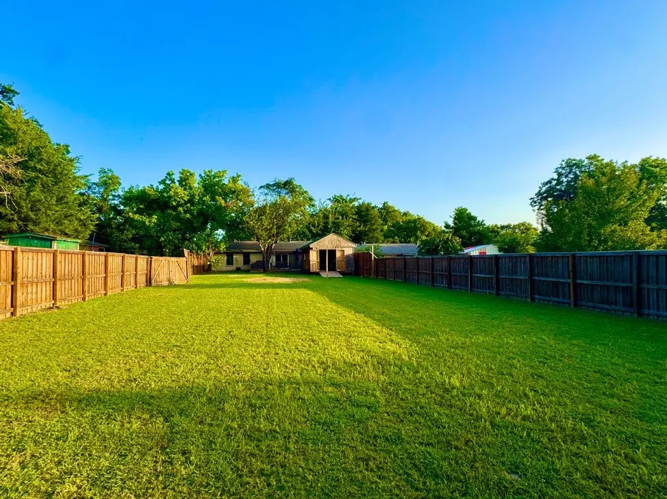 Expansive fenced backyard featuring a large green lawn, mature trees, and a detached outbuilding