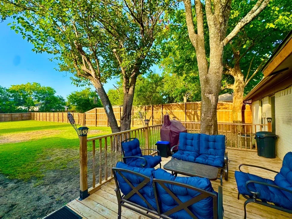 Expansive wooden deck overlooking a spacious lawn, featuring a privacy fence, mature trees, and exterior wall with brick-finish siding