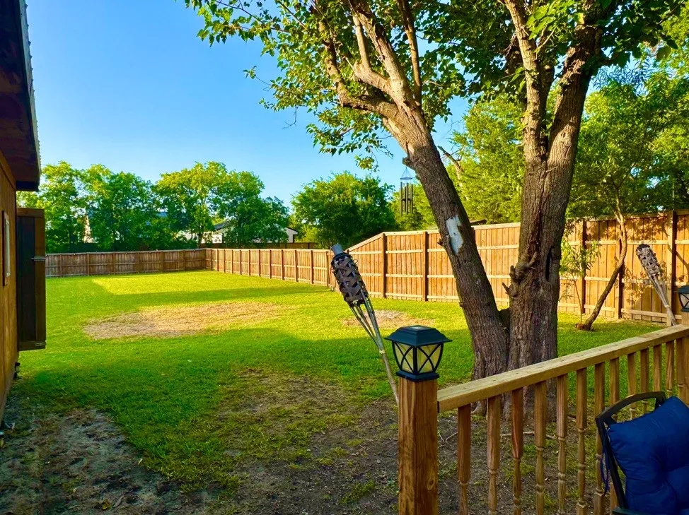 Expansive backyard featuring a wood privacy fence, mature trees, a wooden deck with railing, and a clear blue sky overhead