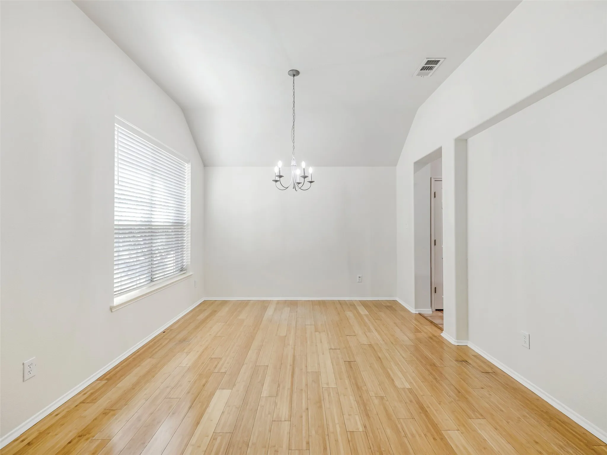 The formal dining room located immediately off the front entry foyer of 414 Dartmoor Dr, Celina, TX. This flexible space features durable light wood-look plank flooring, freshly painted white walls, a vaulted ceiling, and a large window with horizontal blinds for natural light. A decorative six-light candelabra chandelier hangs in the center of the room. This space can also serve as a dedicated home office.