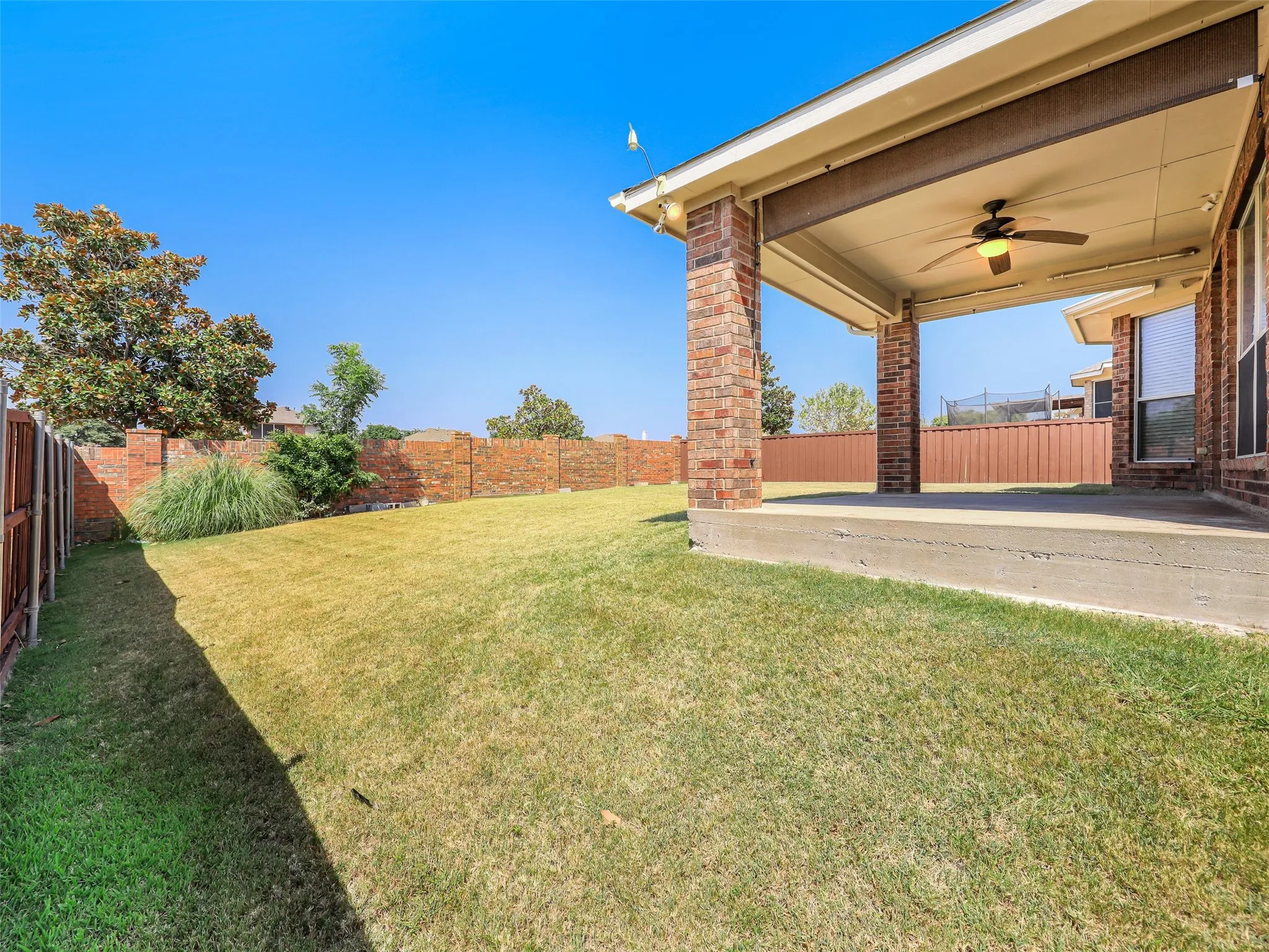 An exterior view of the covered back patio at 414 Dartmoor Dr, Celina, TX. Features classic brick support pillars, an outdoor ceiling fan with light fixture, a concrete slab foundation, a pull-down outdoor privacy shade, and a view looking out onto the spacious grassy backyard enclosed by wooden and brick perimeter fencing.