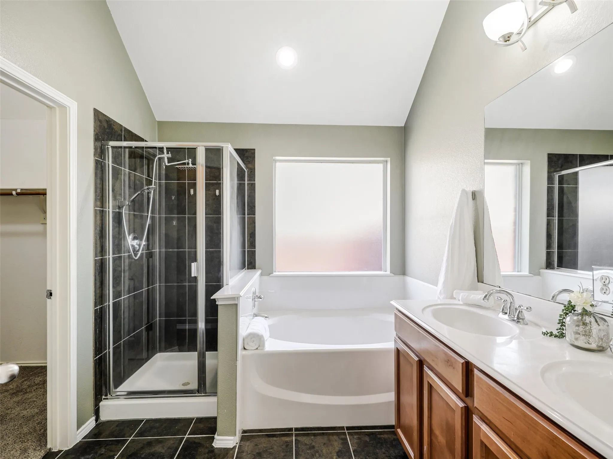 Bathroom featuring a glass-enclosed shower with dark tile surround, a soaking tub with frosted windows, a double vanity with wood cabinetry and dual sinks, and dark tile flooring