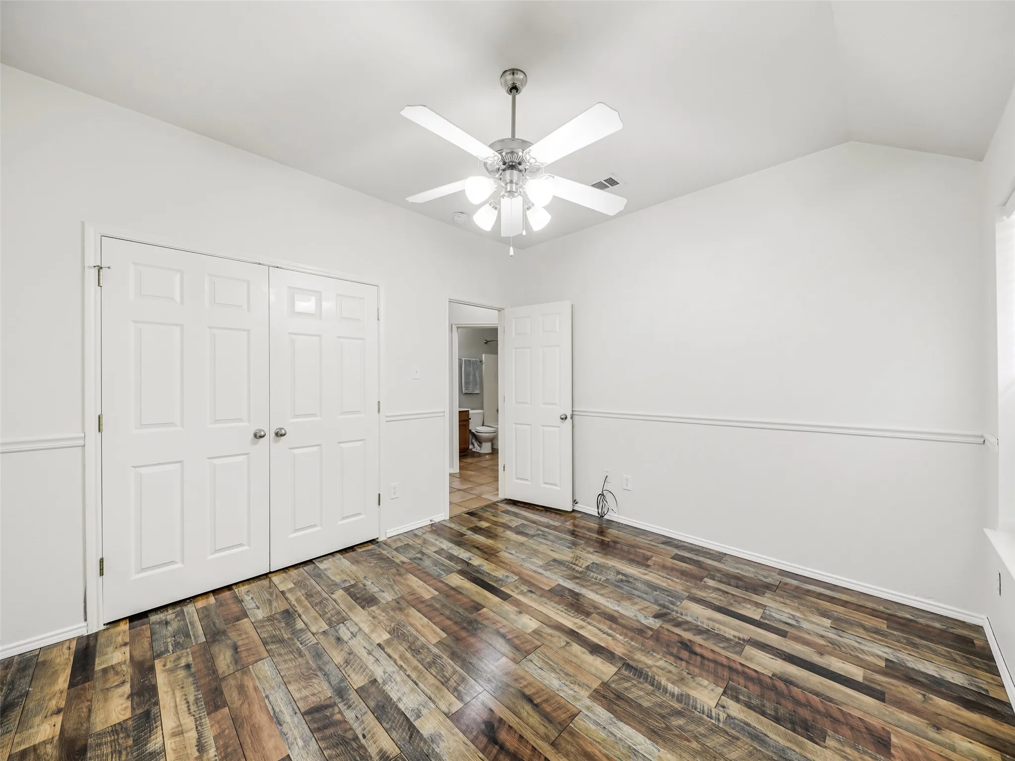 Reverse angle of the secondary bedroom at 414 Dartmoor Dr, Celina, Texas. Showcases the double-door closet, decorative chair rail trim, wood-look plank flooring, white ceiling fan, and direct doorway access to the adjacent guest hallway bathroom.