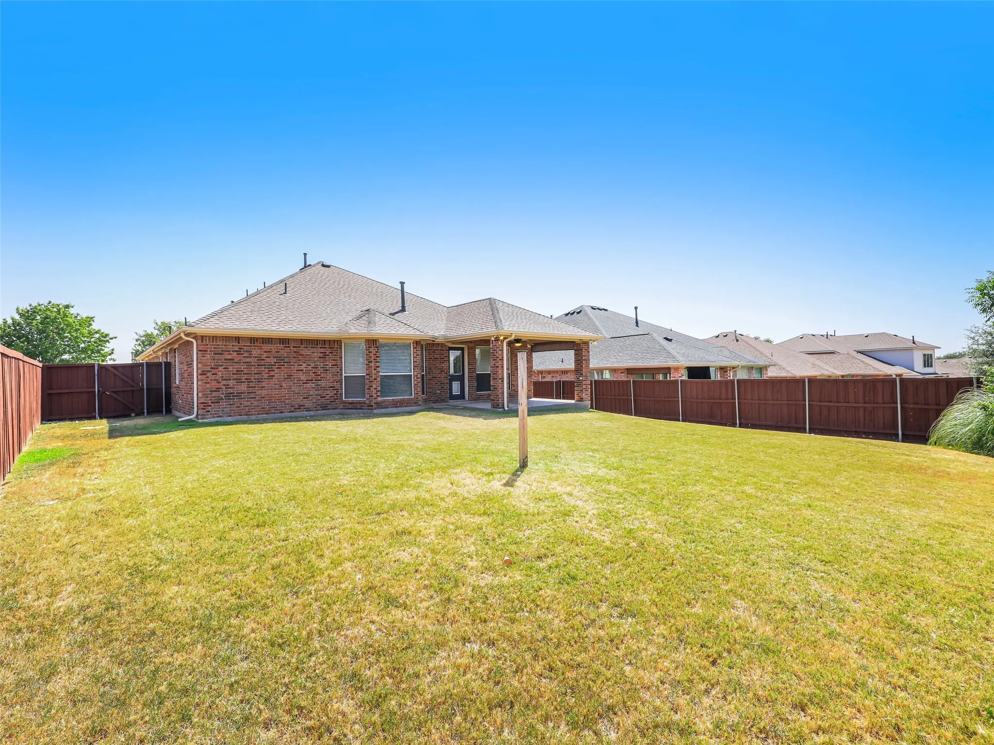 Straight-on perspective of the rear exterior at 414 Dartmoor Dr in Celina's Carter Ranch neighborhood. Showcases the single-story all-brick construction, covered patio extension with ceiling fan, side gate access, and fully enclosed wood privacy fence.