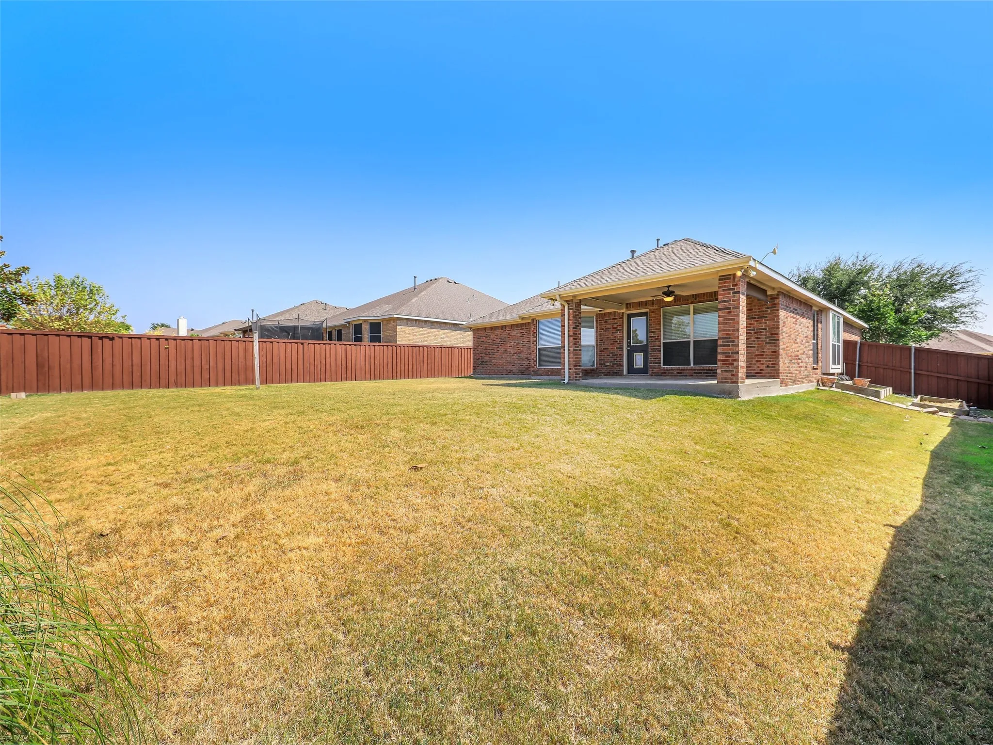 Broad angle overview of the full rear yard at 414 Dartmoor Dr. Shows the all-brick single-story exterior elevation, the extended covered patio area, dark wood privacy fencing along the property line, and an expansive lawn with plenty of room for outdoor entertaining, play, or future additions.