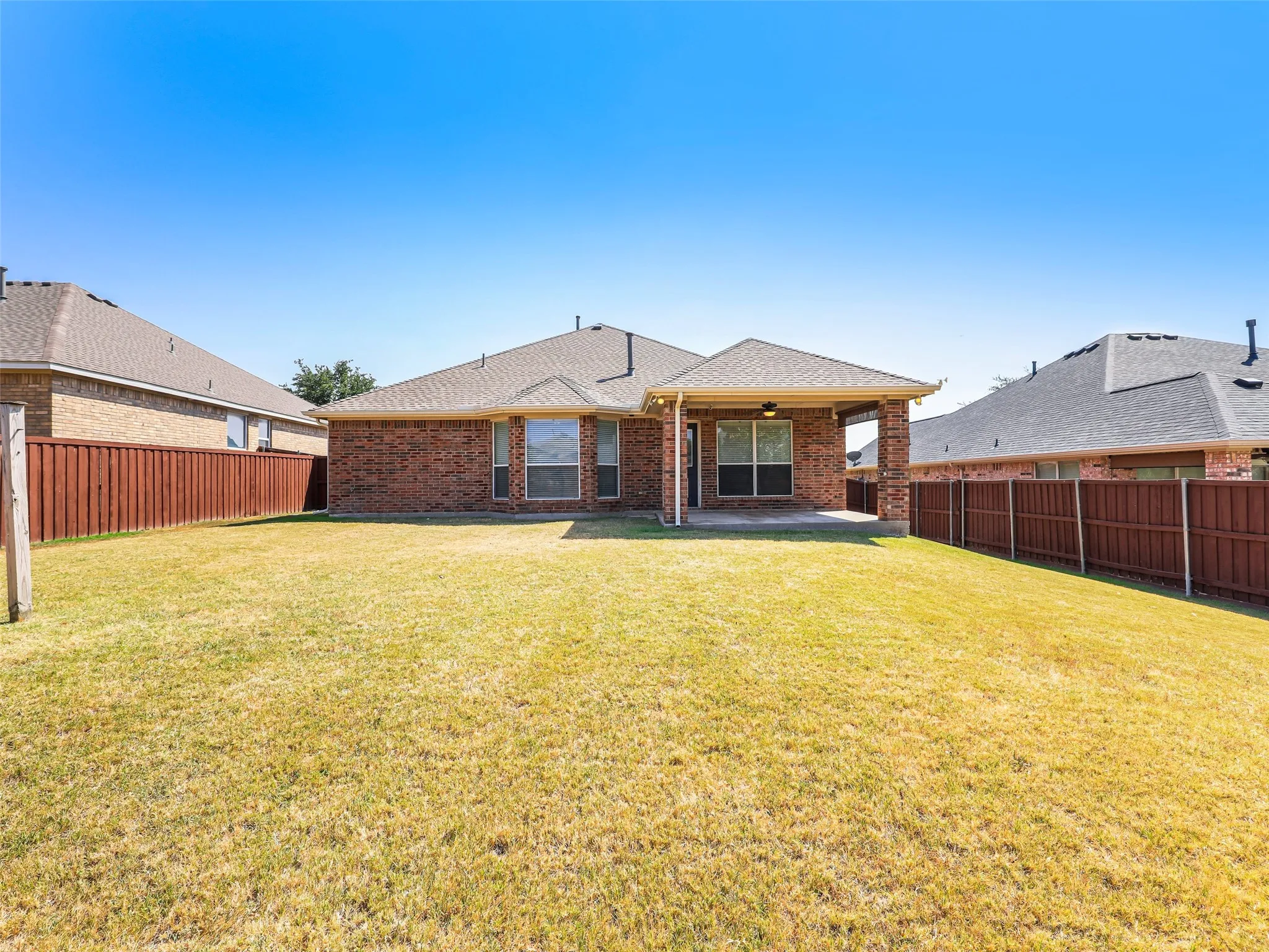 Centered shot from the back fence looking straight at the house at 414 Dartmoor Dr, Celina, TX. Highlights the architectural bay window pop-out from the primary suite, the covered patio structure, composite shingle rooflines, and a vast open lawn under clear blue Texas skies.