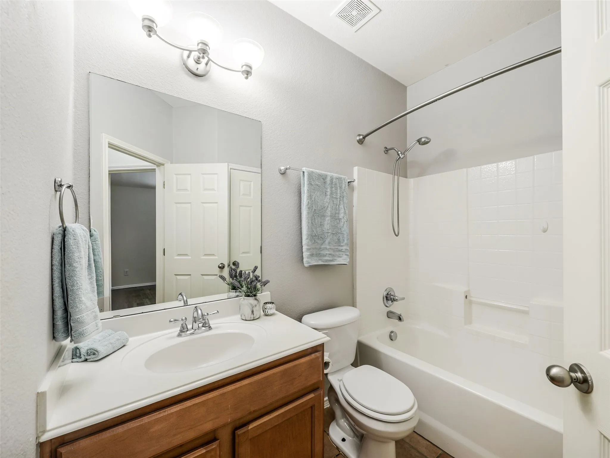 Full guest bathroom located at 414 Dartmoor Dr in Carter Ranch, Celina, TX. Includes a wood single-sink vanity with white countertop, 3-light vanity light fixture, large mirror, standard toilet, shower/tub combination with white tile surround, and neutral tile flooring.