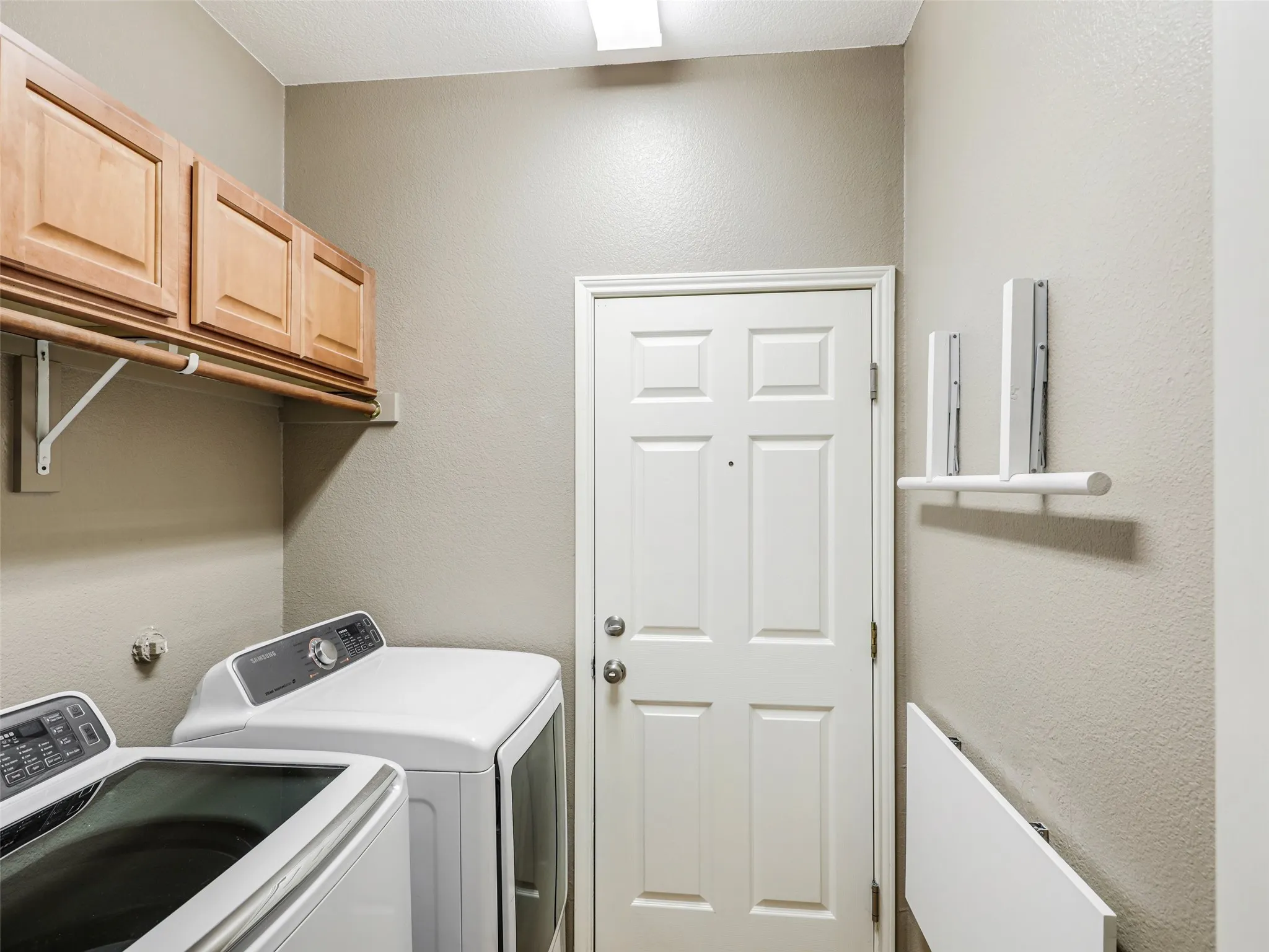 Dedicated interior laundry utility room at 414 Dartmoor Dr, Celina, Texas. Features washer and dryer hookups with Samsung appliances, built-in upper wood storage cabinetry, a clothing hanging rod, wall-mounted folding shelf racks, and direct interior access to the garage through a solid entry door.