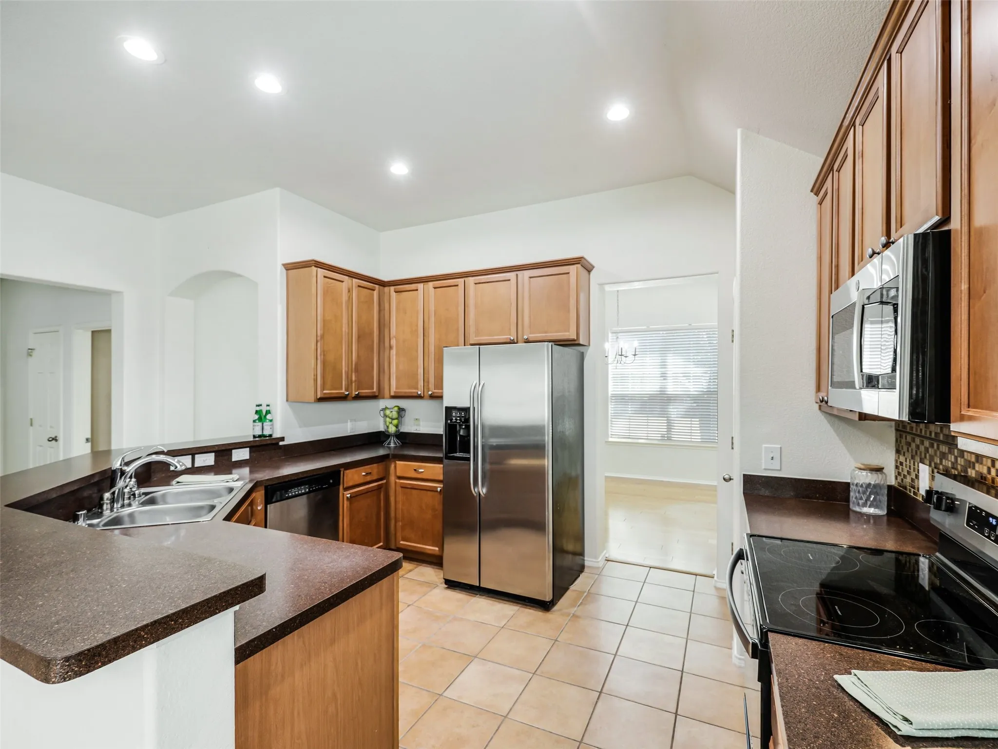 View of the functional kitchen layout at 414 Dartmoor Dr, Celina, TX. This kitchen includes side-by-side stainless steel refrigeration, an electric smooth-top range with an built-in microwave above, wood cabinets, dark counters, and a mosaic glass-tile backsplash. Large ceramic floor tiles extend throughout the kitchen and main pathways.