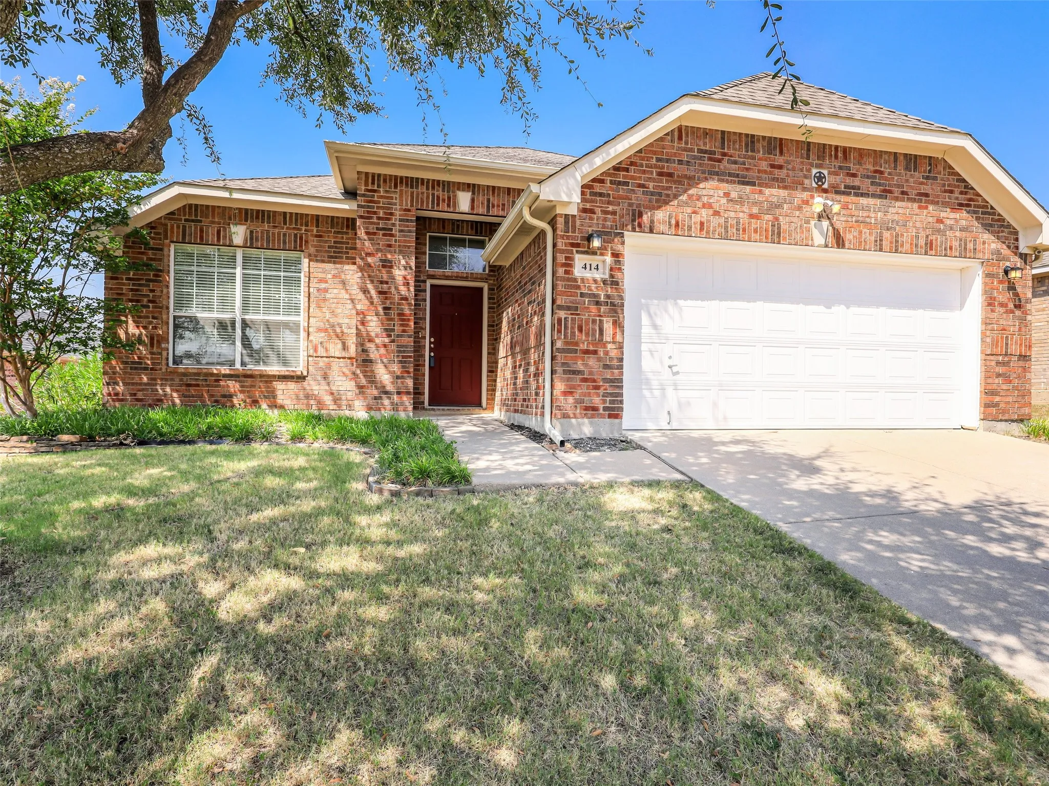 A wide-angle street view showcasing the curb appeal of 414 Dartmoor Dr in Celina's Carter Ranch community. This single-story brick home has a large front lawn, an expansive concrete driveway leading to the 2-car garage, and established landscaping. Mature trees provide significant shade to the property. The roof features architectural shingles and includes a decorative star emblem near the peak.