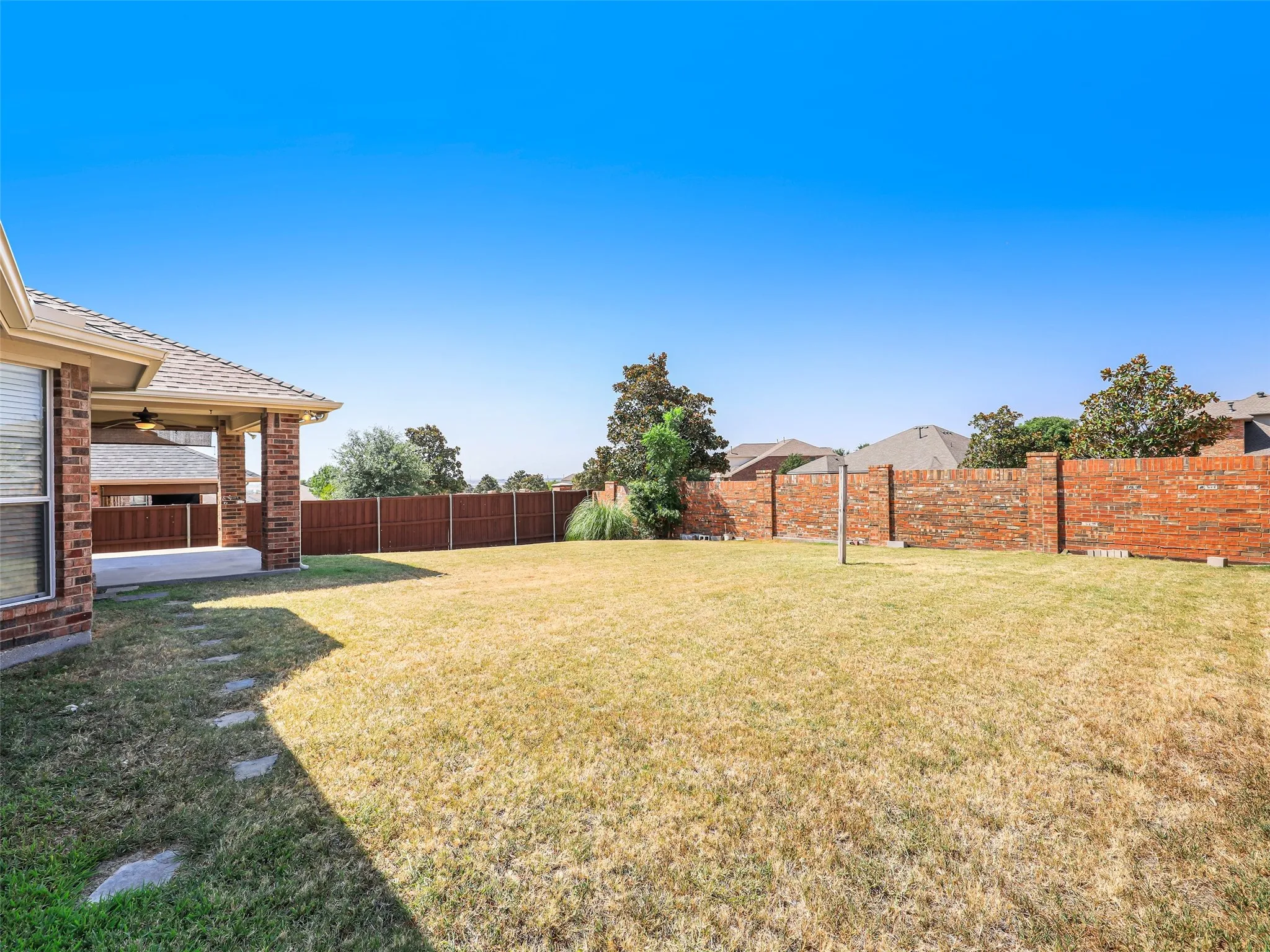Angle showing the expansive green lawn space in the backyard of 414 Dartmoor Dr in Carter Ranch, Celina, Texas. Highlights include stepping stone pathways along the side, a combination of wood privacy fencing and a decorative red brick perimeter wall, and mature landscaping accents.
