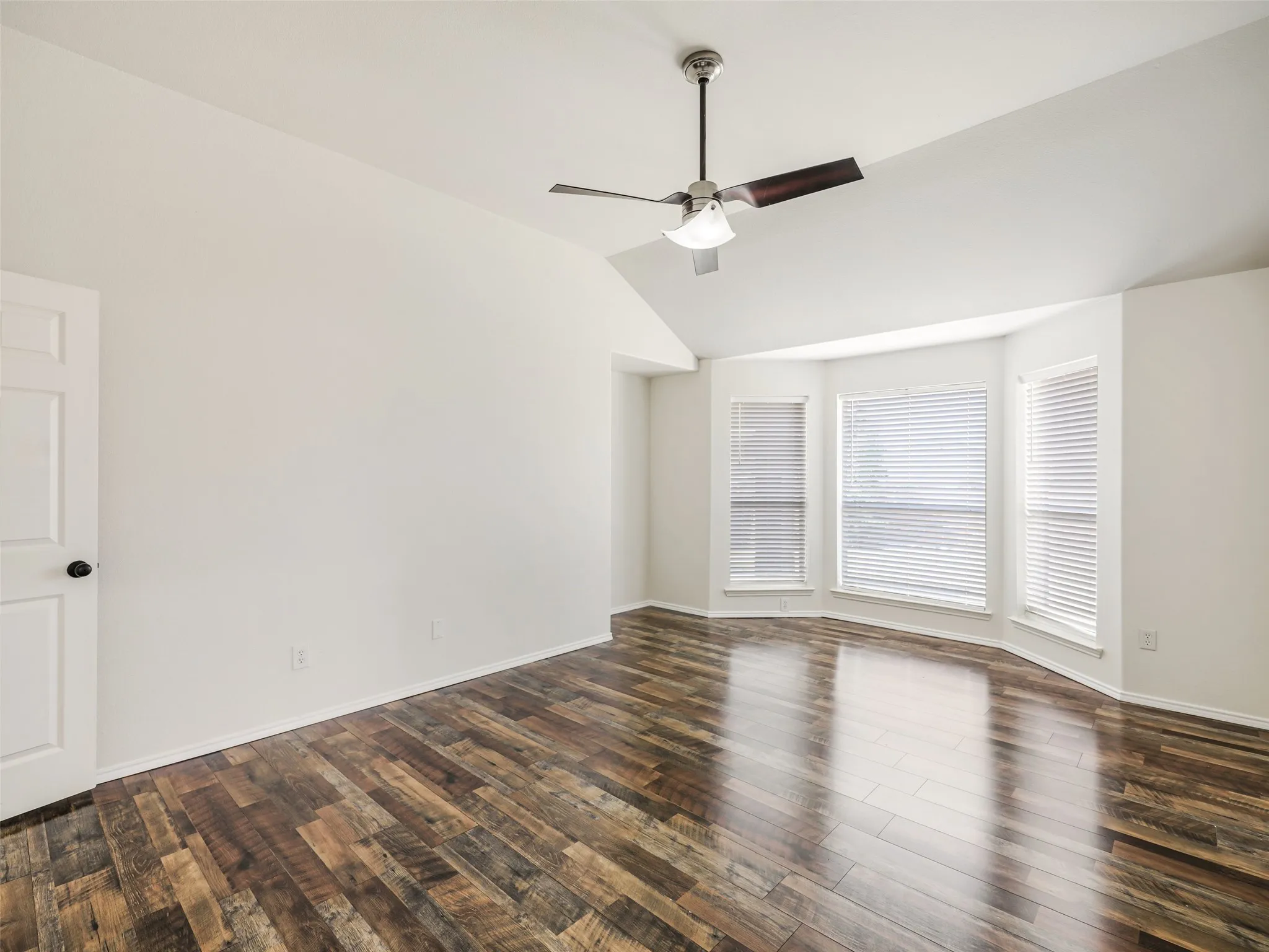 Interior view of the primary bedroom at 414 Dartmoor Dr, Celina, TX, showcasing the layout toward the ensuite bath and hallway entry. Features high vaulted ceilings, modern dark-wood multi-tone plank flooring, neutral painted walls, and a 5-blade ceiling fan with an integrated light fixture.