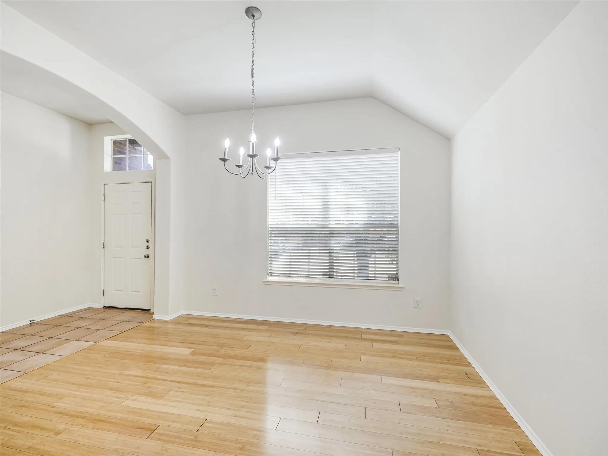 An interior view looking from the main entryway foyer toward the formal dining area. The perspective highlights the architectural transition with a curved archway that connects the spaces. The flooring changes from large ceramic tile in the foyer to light wood-look plank flooring in the dining room. The room includes a large window with blinds and a simple six-light silver-tone chandelier.