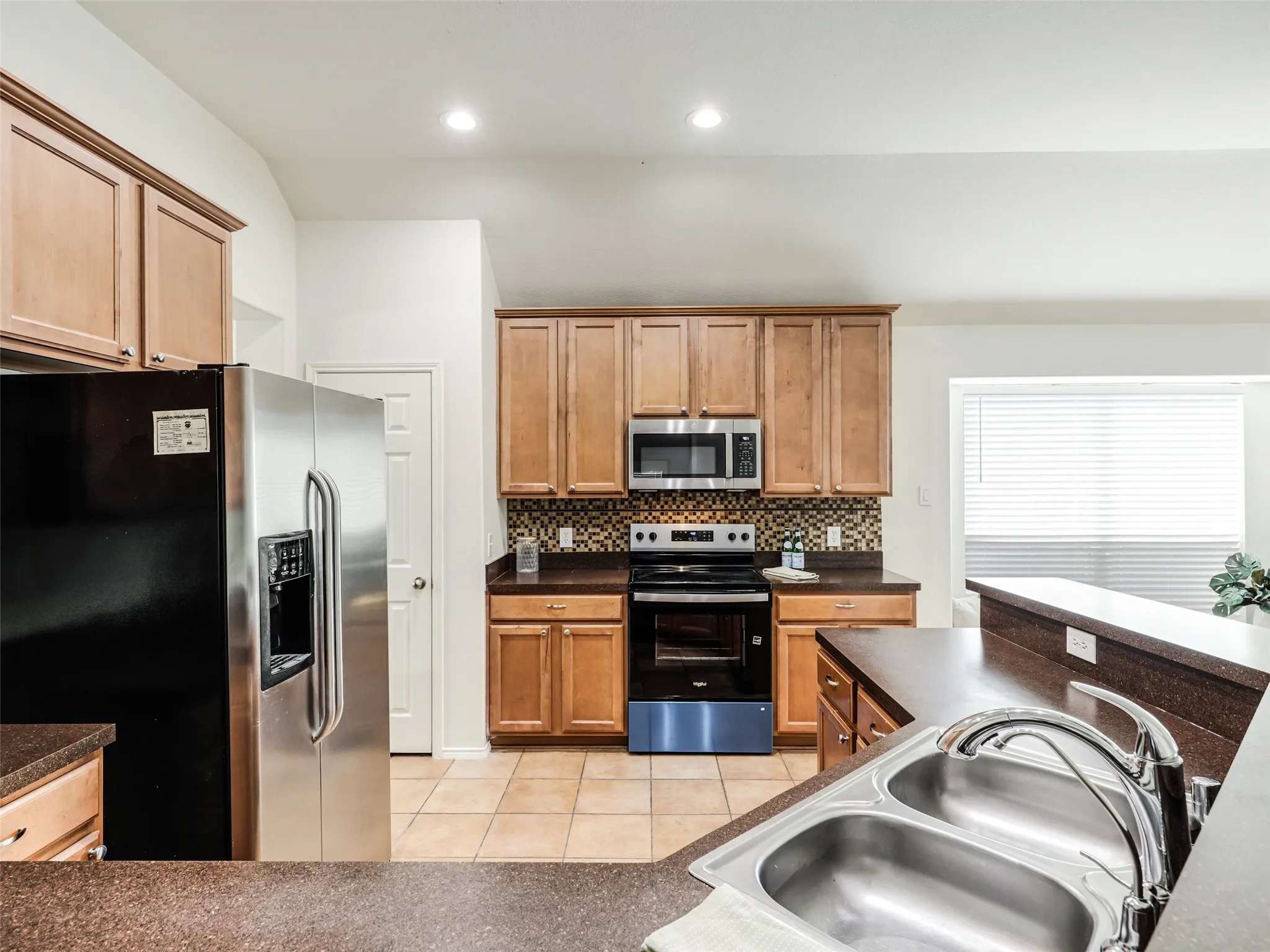 Detailed perspective of the kitchen prep area at 414 Dartmoor Dr, Celina, Texas. Features a double-bowl stainless steel sink with goose-neck faucet set into dark countertops, a smooth-top range with built-in microwave, a mosaic tile backsplash, and a adjacent walk-in pantry door for food storage.