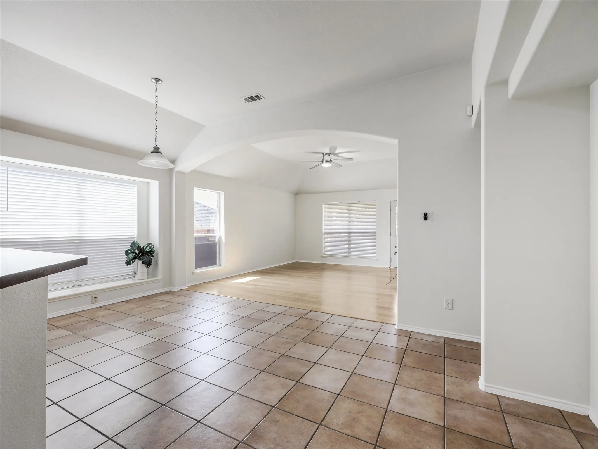 An interior view showing the transition between the tiled eat-in dining nook and the spacious main family room at 414 Dartmoor Dr in Carter Ranch. The space features large neutral ceramic floor tiles leading into warm wood-look plank flooring under a grand architectural archway. A modern silver ceiling fan anchors the living space.