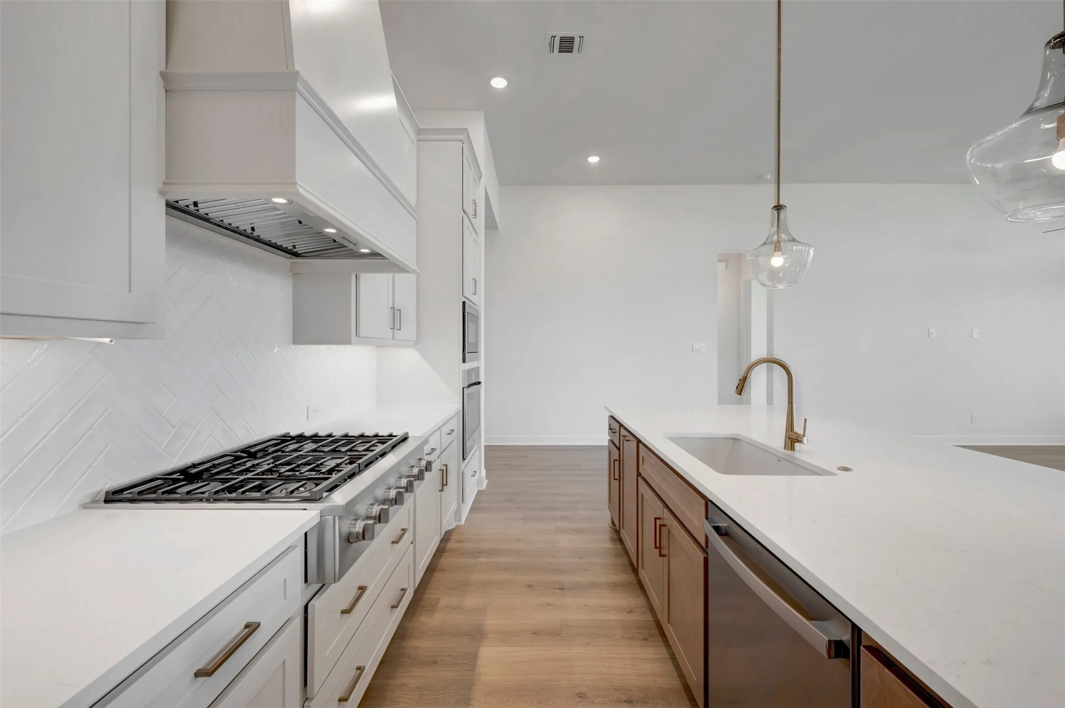 Gourmet kitchen featuring a built-in gas range, white herringbone tile backsplash, white shaker cabinetry, an expansive island with a built-in sink, and pendant lighting