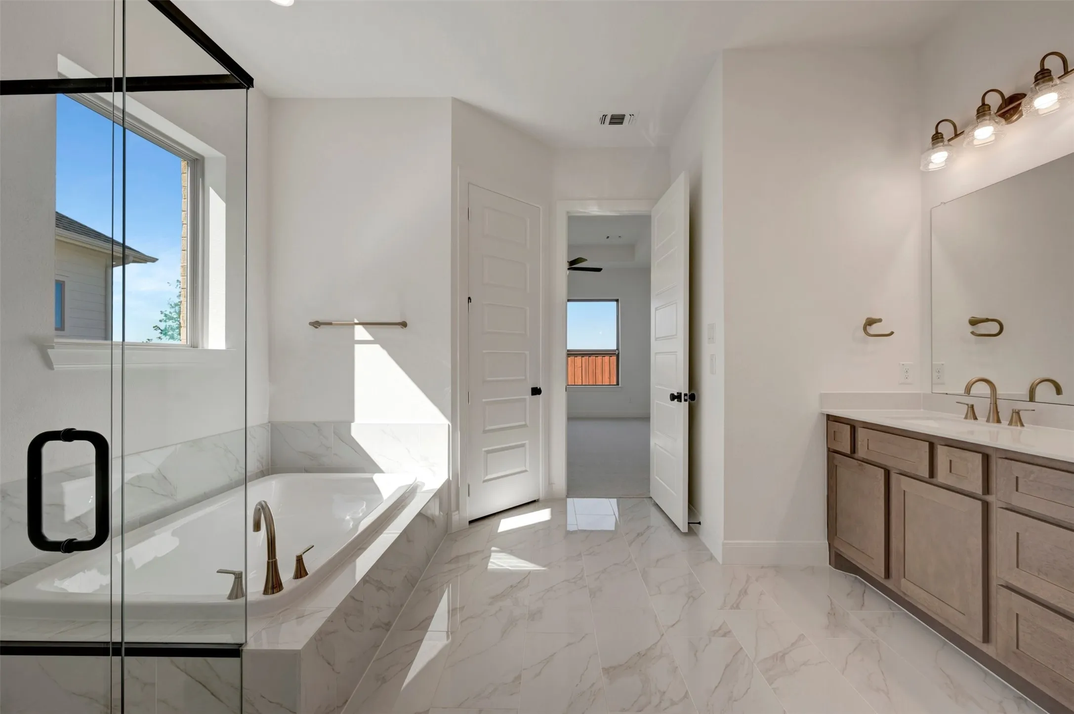 White tile flooring, built-in bathtub with tile surround, glass-enclosed shower, dual vanity with undermount sinks, and wall-mounted light fixture