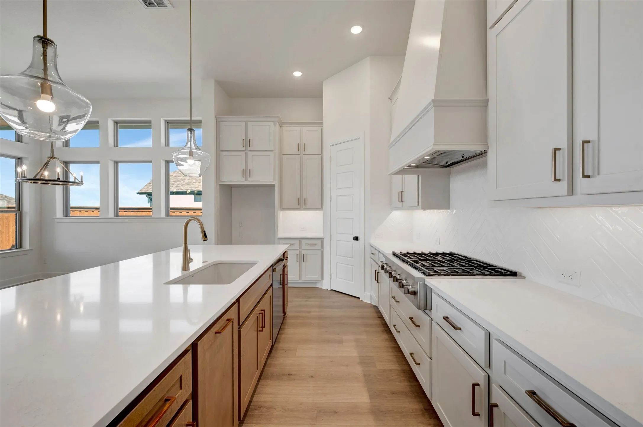 Kitchen featuring an expansive island with an undermount sink, gas cooktop with a custom range hood, white shaker-style cabinetry, and wood-finish flooring