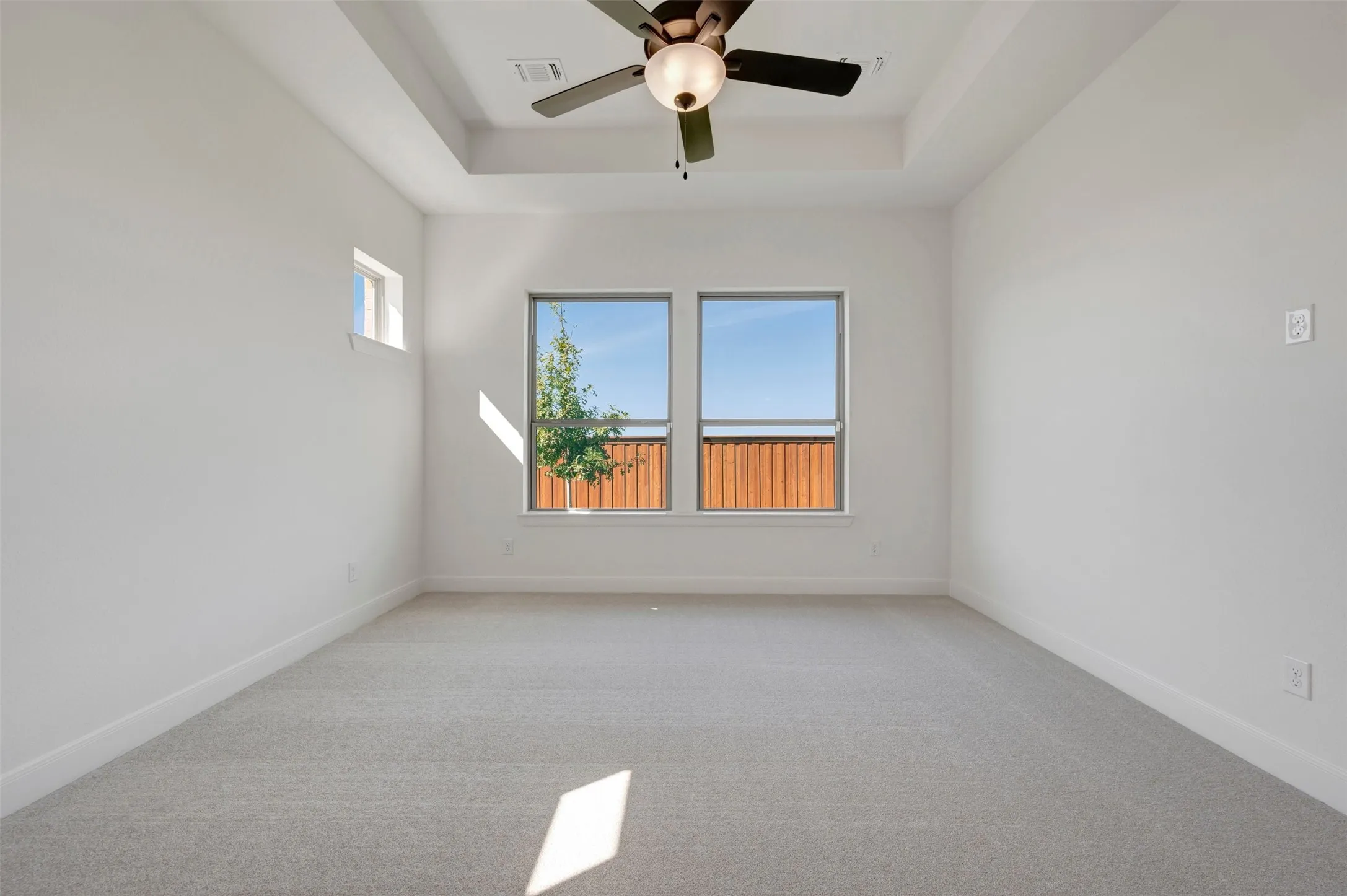 Tray ceiling with a ceiling fan, two large windows, a high window, and light gray carpeting