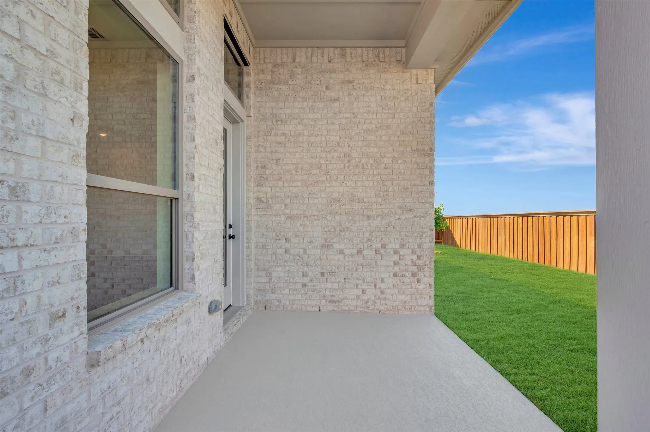 Covered patio with a concrete surface, white brick exterior, large window, and a privacy fence bordering a manicured lawn