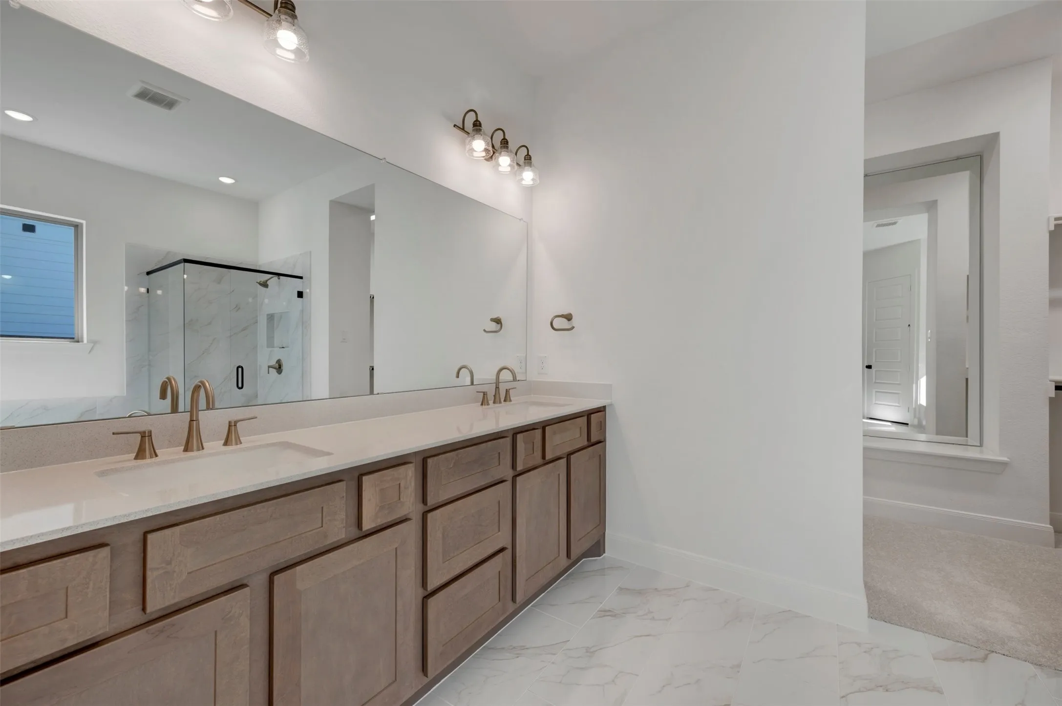 Dual vanity with brushed gold fixtures, light wood-finish cabinetry, white stone-finish countertop, and a full-width mirror