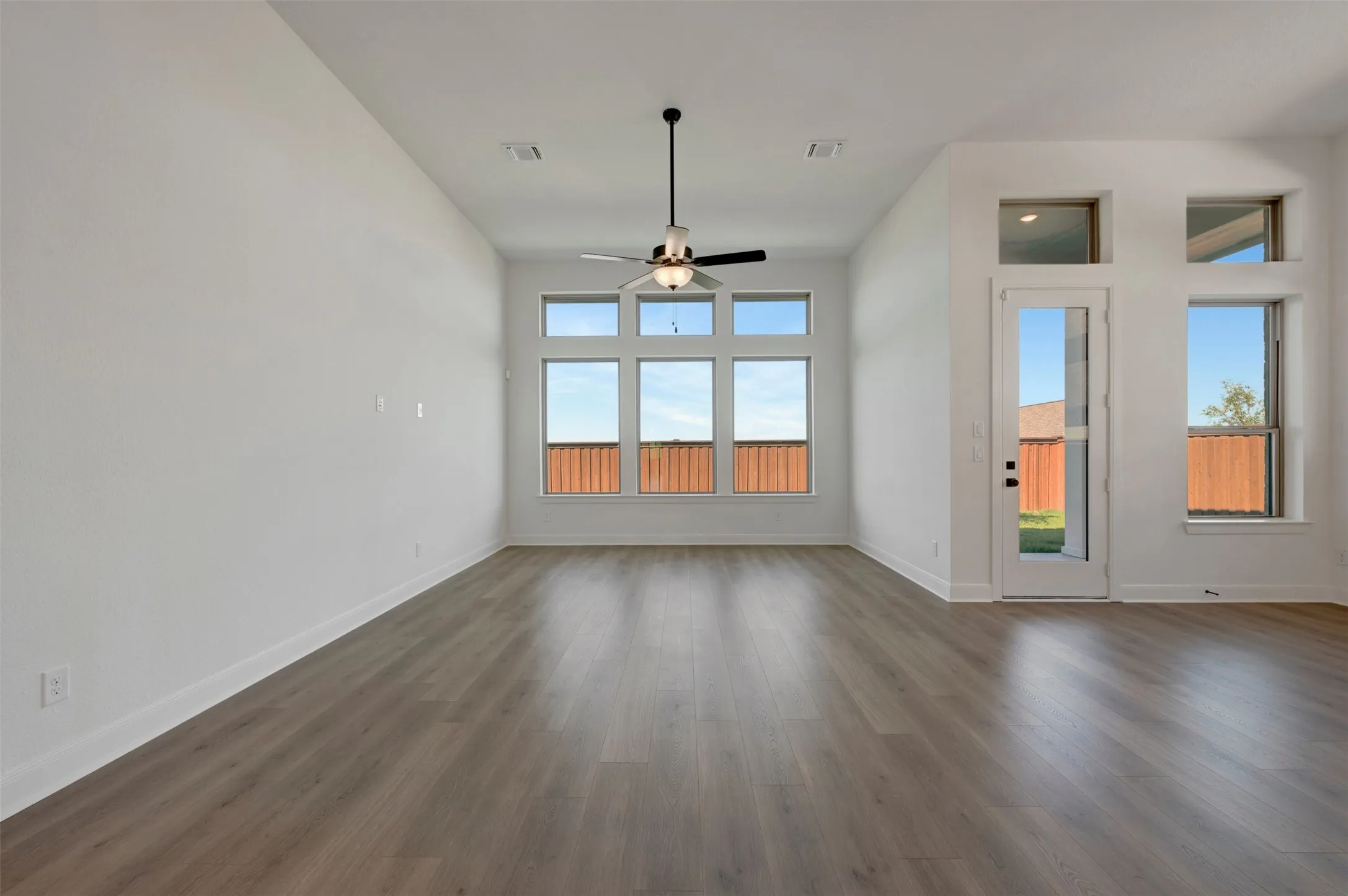 Spacious room with high ceilings, wood-finish flooring, abundant natural light from multiple windows, and a ceiling fan