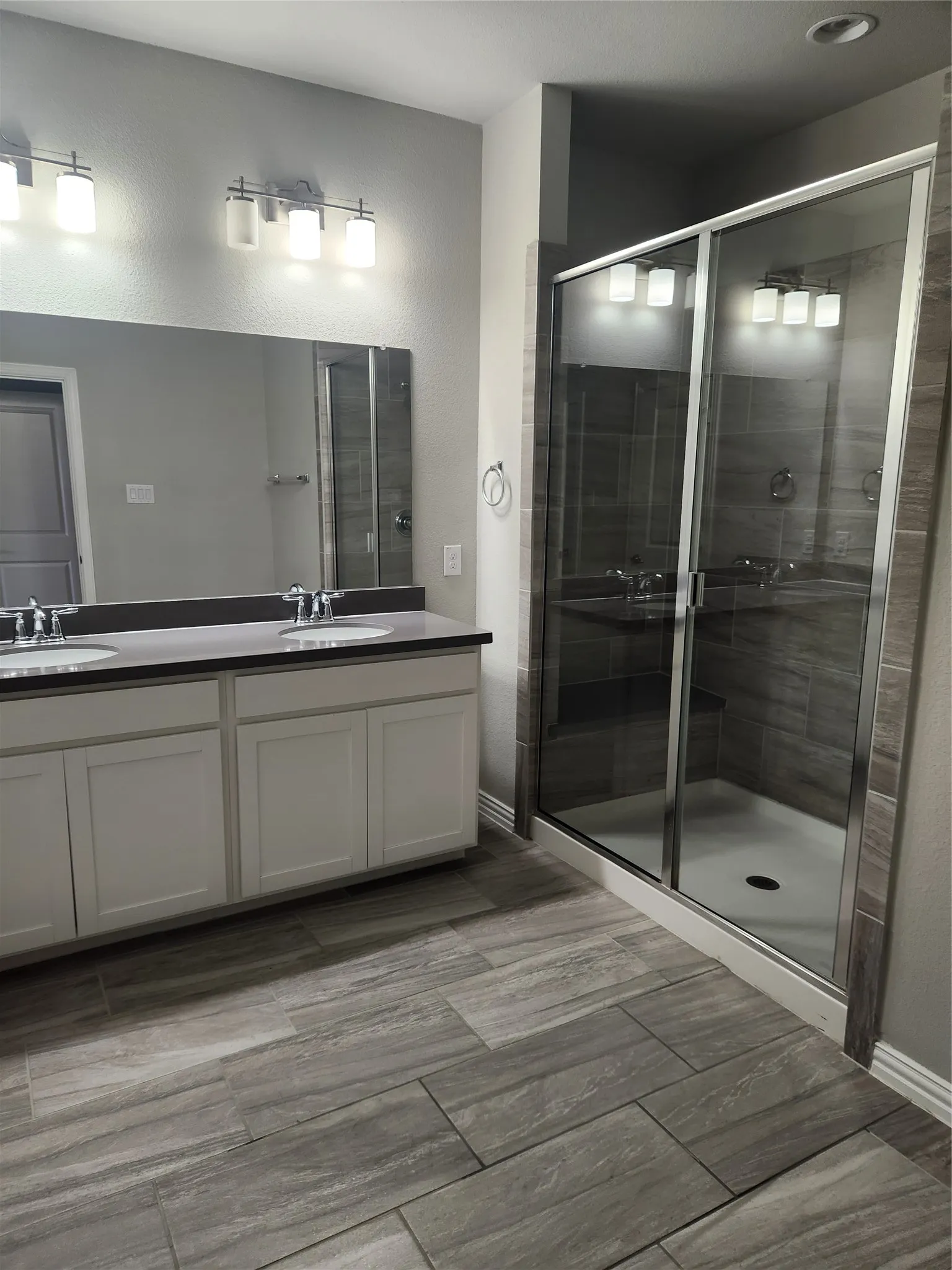Spacious bathroom featuring a glass-enclosed shower with tiled walls, dual vanity with white cabinetry, black countertops, and a full-width mirror