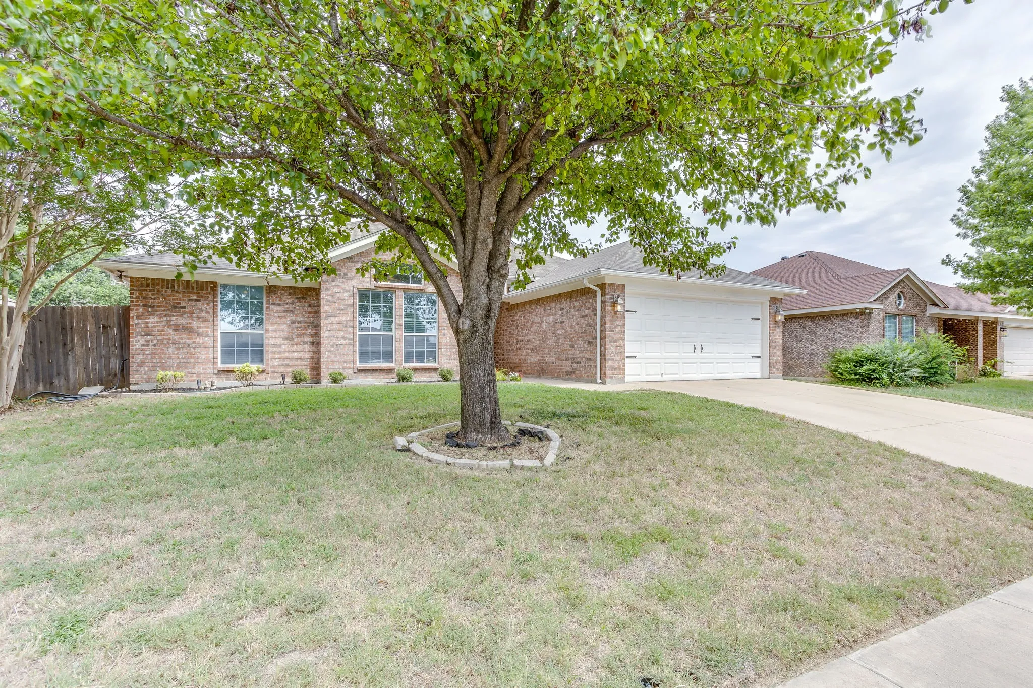 Front of house features red and tan multi-colored brick, concrete slab driveway, and mature shade tree with a stone border.