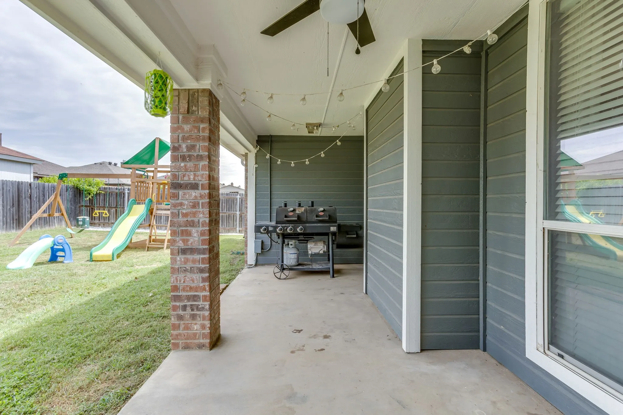 A spacious covered patio connected to the back yard featuring dark horizontal siding, white flat-panel ceiling and a multi-toned brick column.