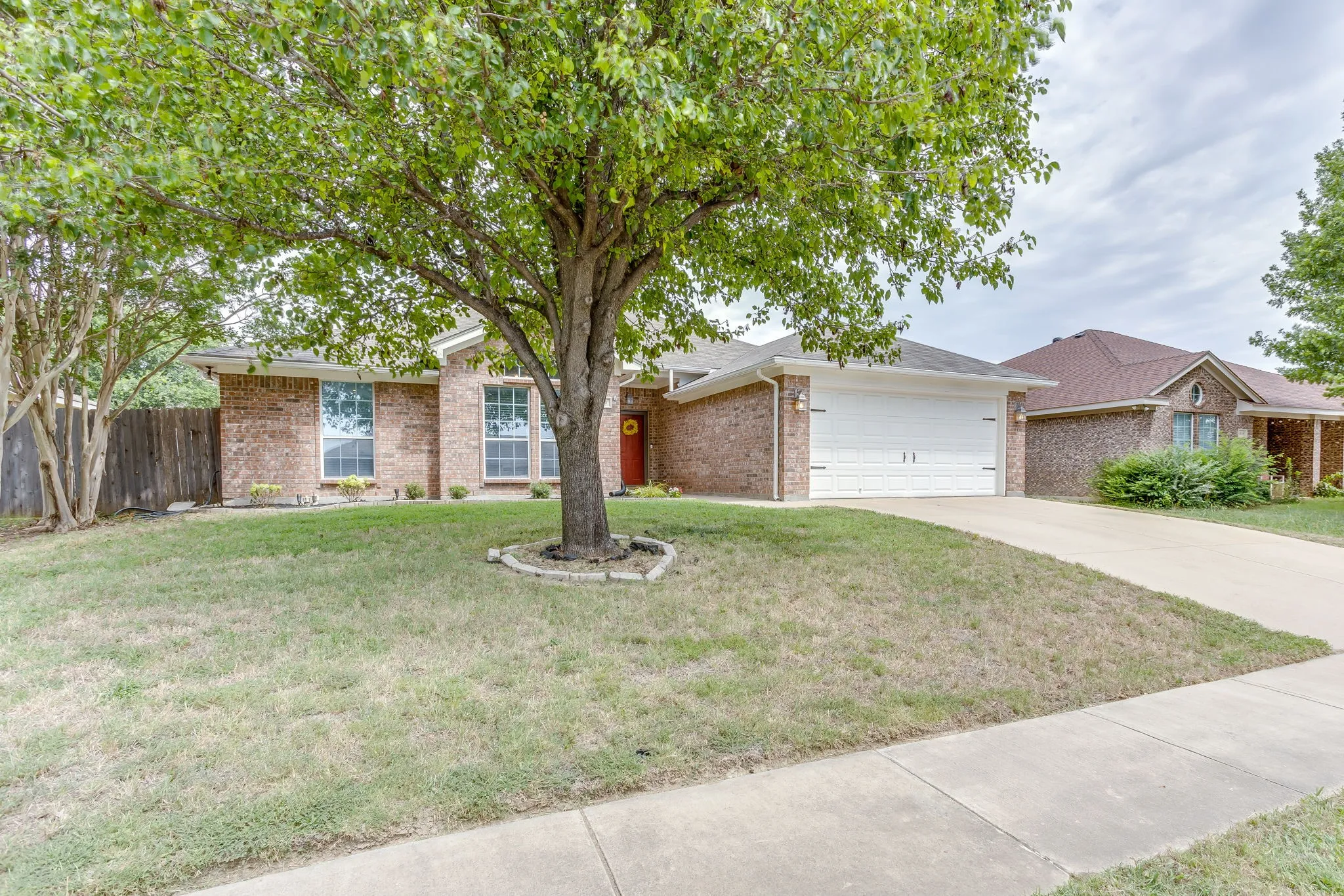 Front of house features red and tan multi-colored brick, concrete slab driveway, and mature shade tree with a stone border.