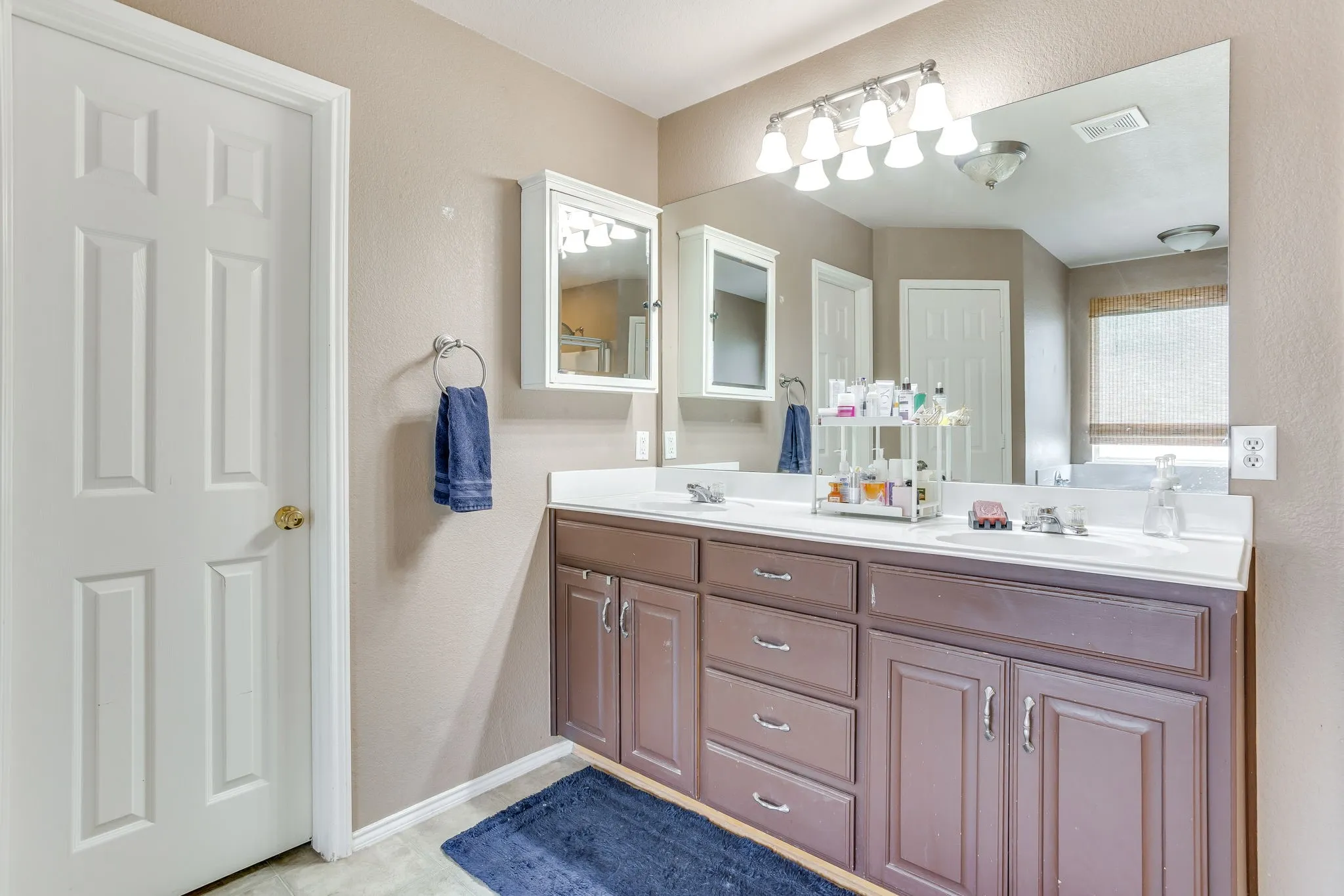 Primary bathroom featuring dual vanity sinks with dark brown wood tone and traditional raised-panel design, and a multi-bulb vanity light fixture with frosted white bell glass shades.
