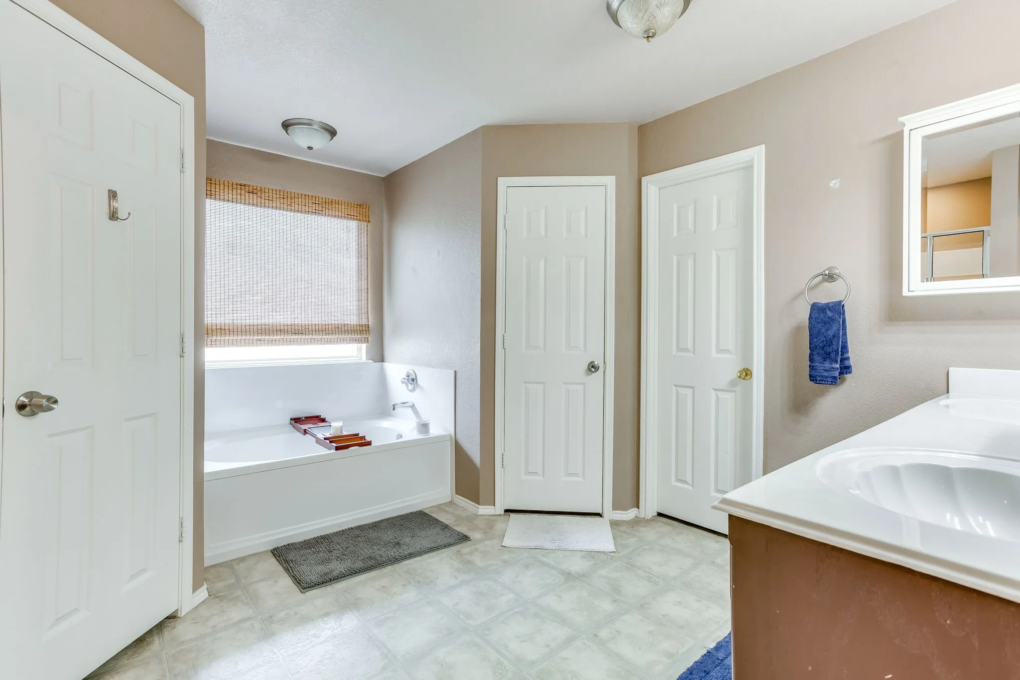 Primary bath featuring large-format rectangular tile-style flooring in a light gray stone pattern, white soaker tub with large window for natural lighting, and classic style 6 panel doors.