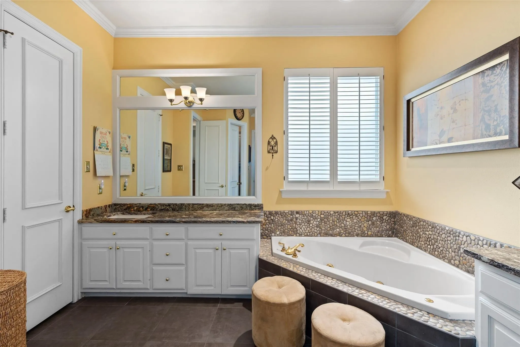Bathroom featuring a corner soaking tub with pebble tile surround, white plantation shutters, a white vanity with integrated cabinetry, and a stone-finish countertop