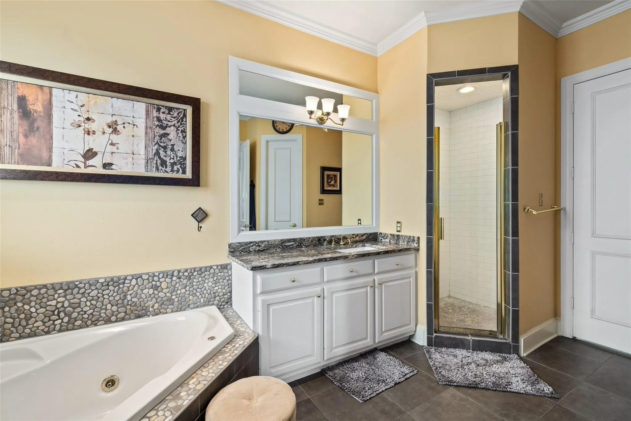 Bathroom featuring a corner soaking tub with pebble tile surround, a single vanity with stone countertop, a framed mirror, and a glass-enclosed shower with subway tile